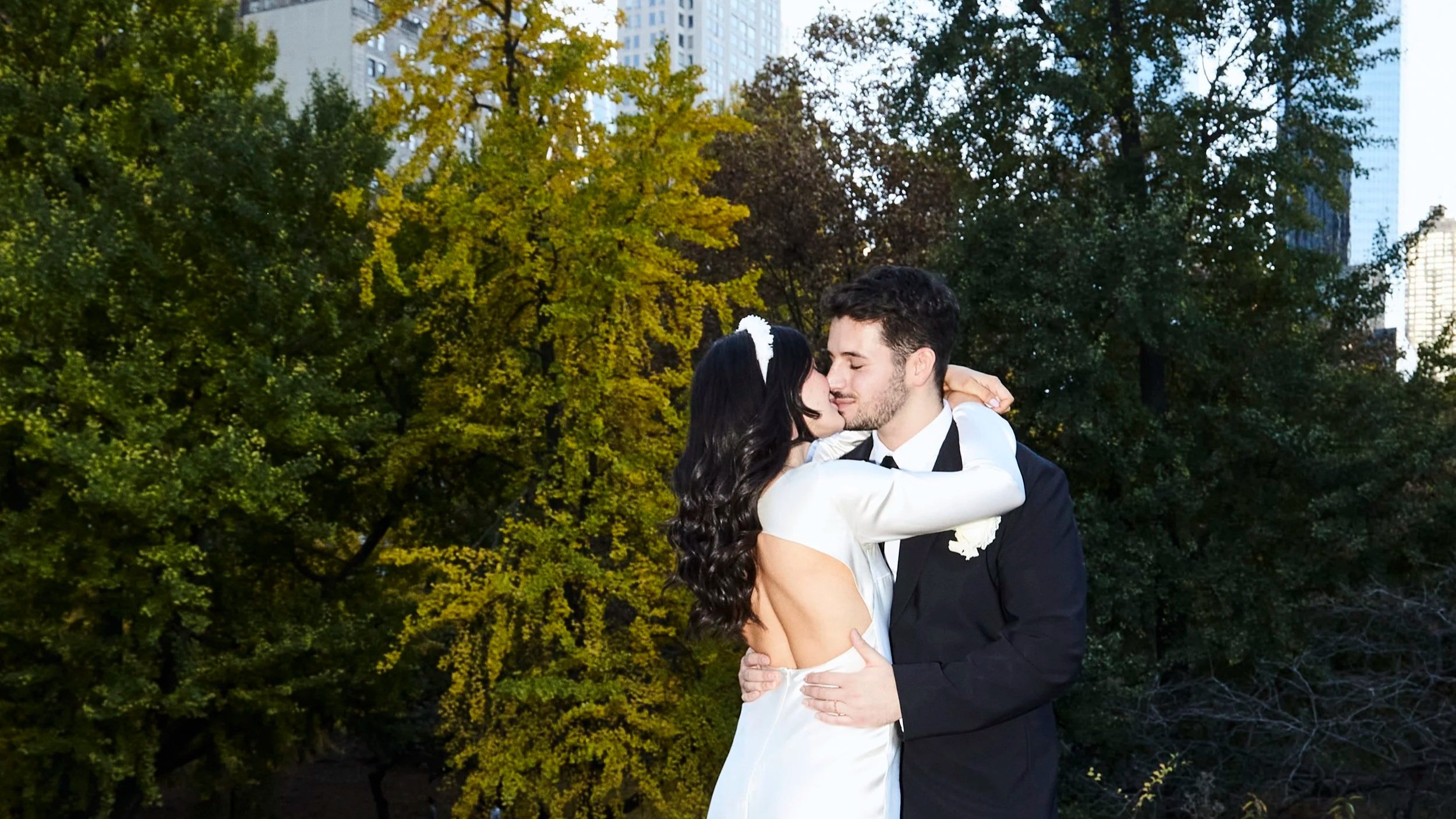 A couple is hugging and kissing in a park with green trees and tall buildings in the background.