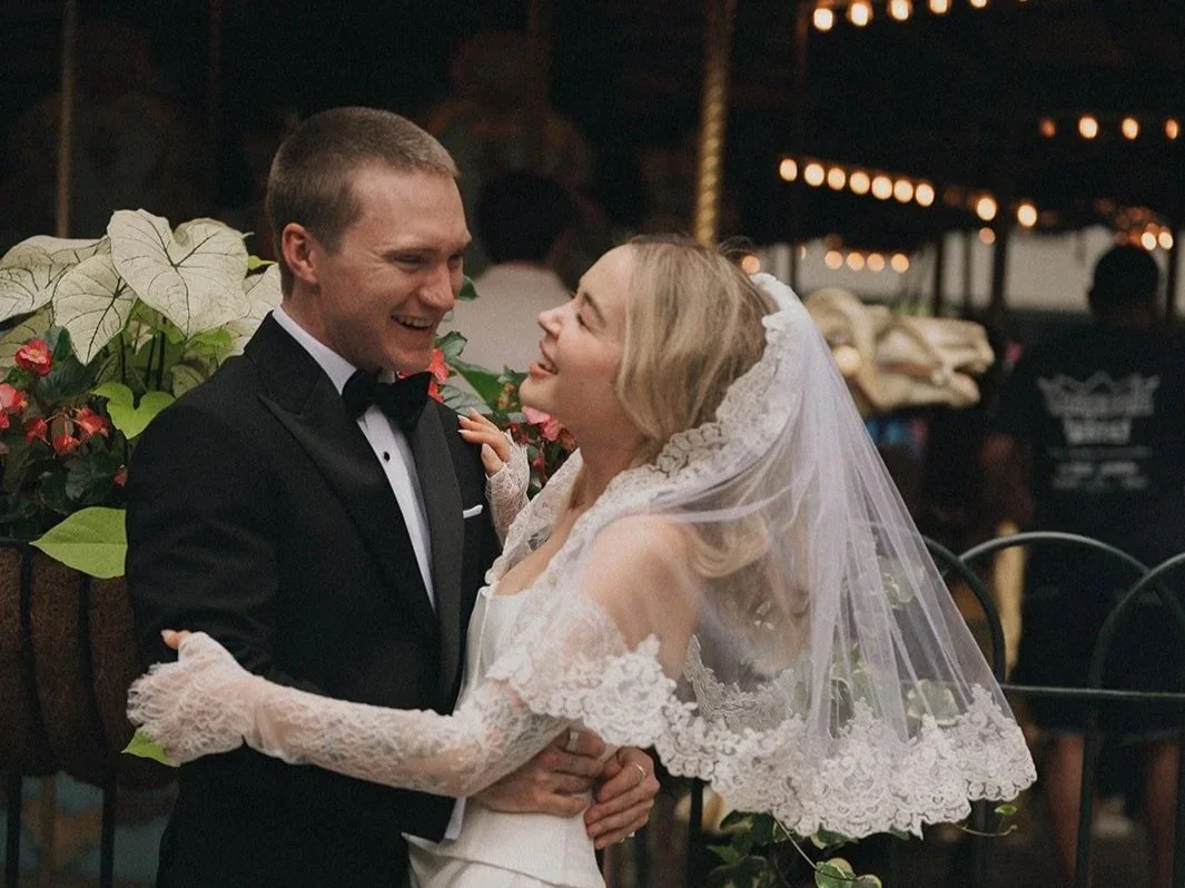 A bride and groom sharing a joyful moment, standing close together, with the bride wearing a lace wedding dress and veil, and the groom in a black tuxedo, in a festive setting with string lights in the background.