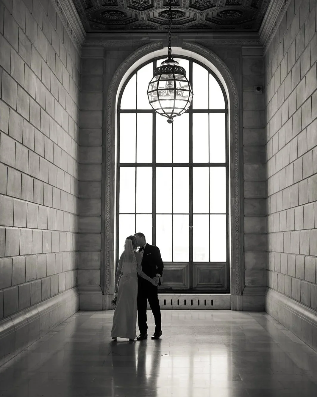 A black-and-white photo of a bride and groom standing close together, holding hands, inside a grand hall with large arched window and ornate ceiling, illuminated by a hanging chandelier.