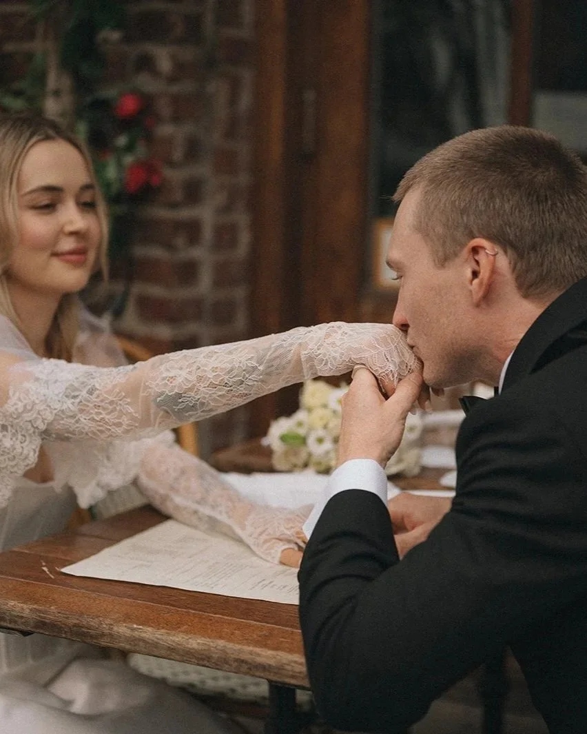A bride and groom at a wedding ceremony; the groom is kissing the bride's hand while she gently touches his forehead. The bride is wearing a lace dress, and the groom is in a black tuxedo. They are seated at a wooden table with a flower arrangement a
