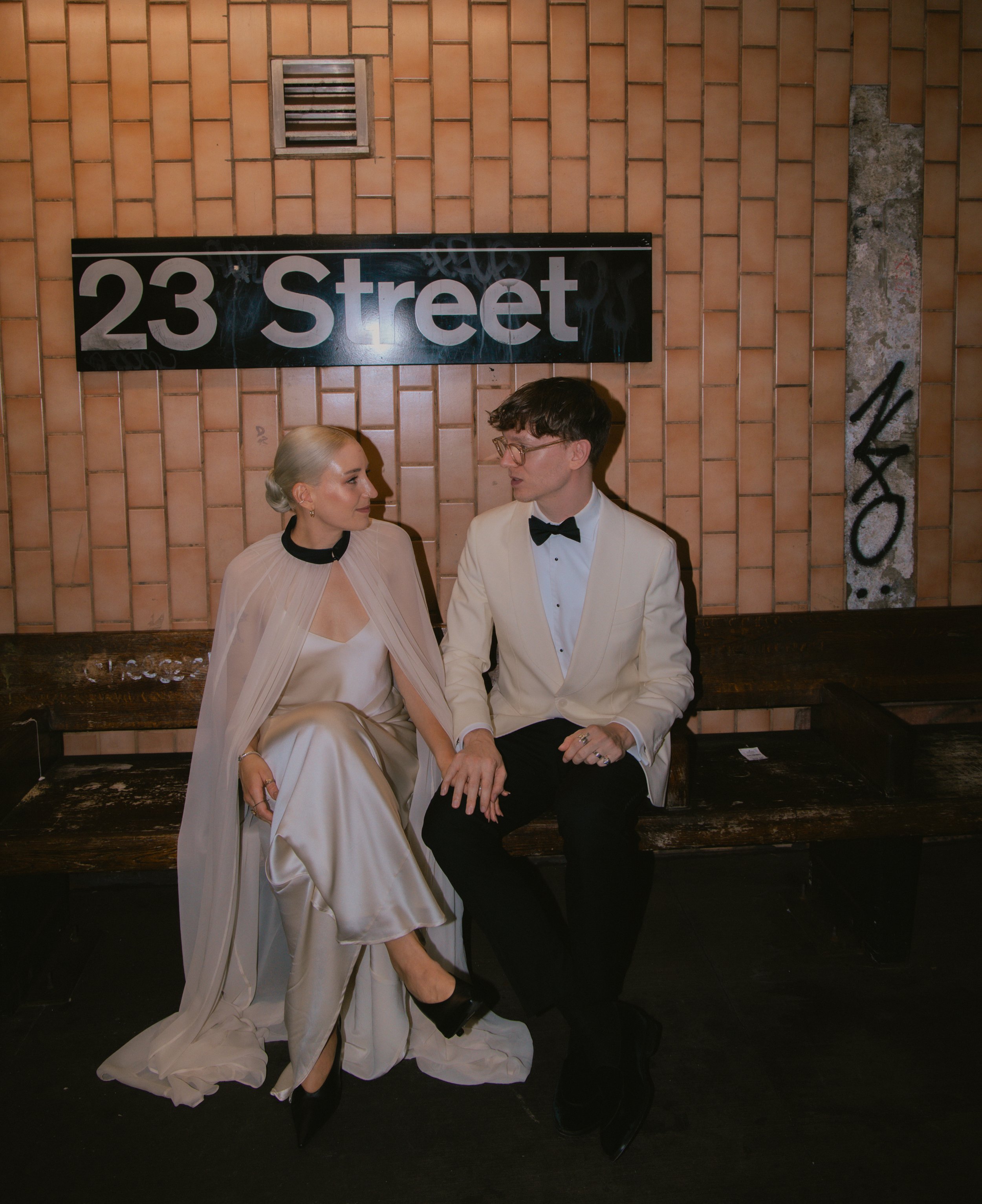 Bride and groom sitting on a bench with the tile wall behind them and '23 Street' sign in the subway station.