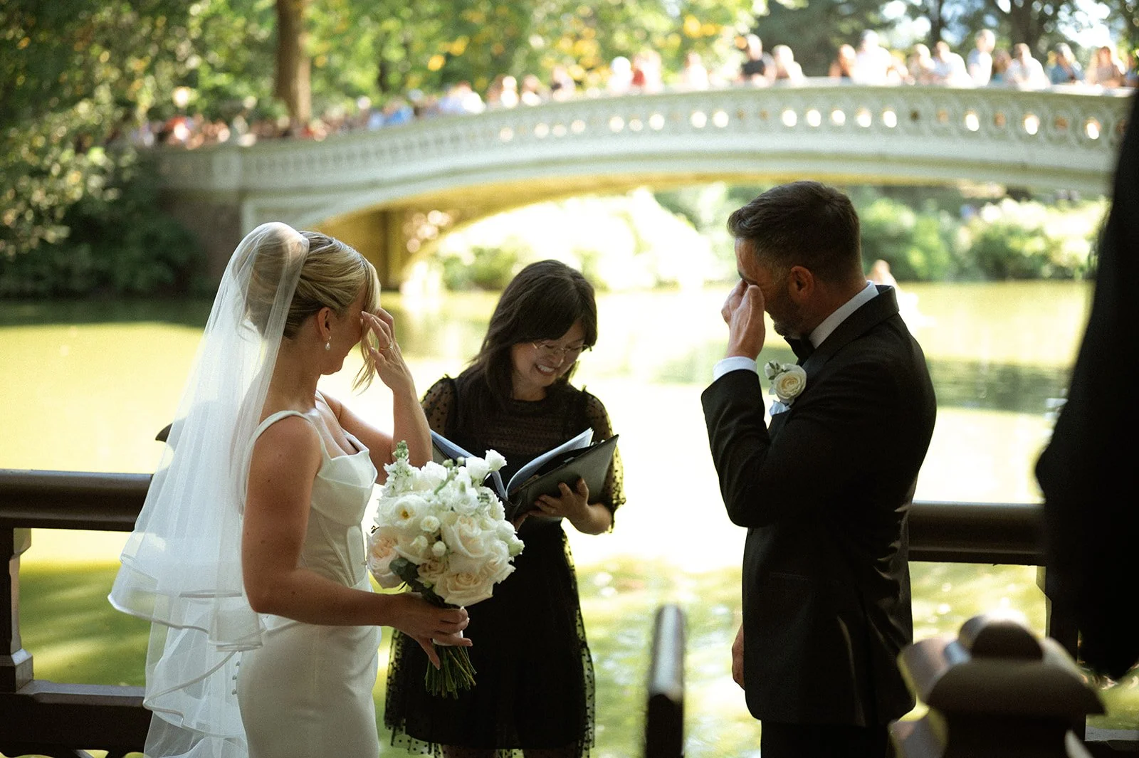 Bride and groom crying tears of joy in front of Bow Bridge in Central Park, NYC while their Cakewalk officiant reads their wedding ceremony speech in front of the water.