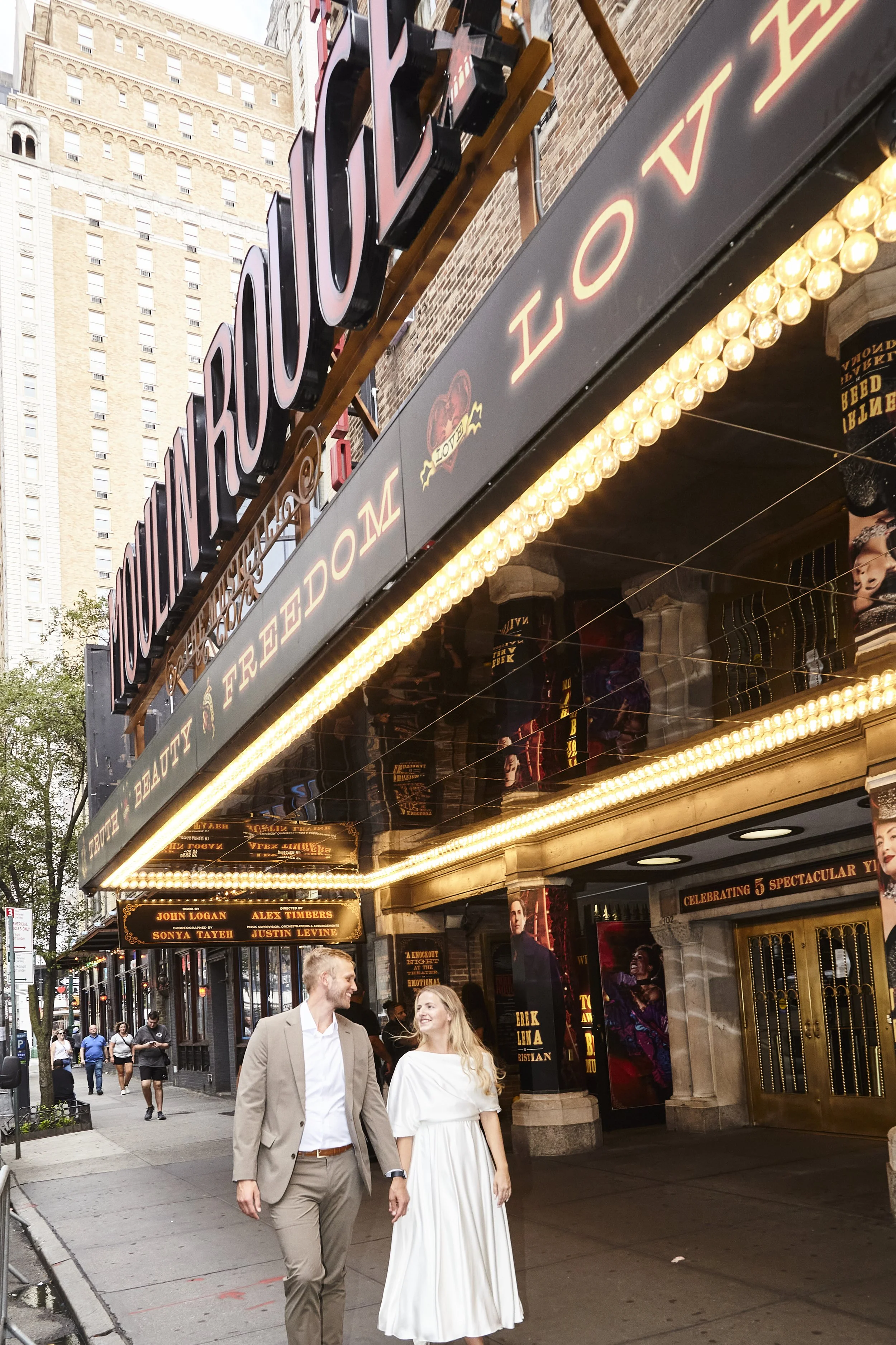 Bride and groom walking in front of the Al Hirschfeld Theatre, where the marquee is lit up and reads Moulin Rouge!