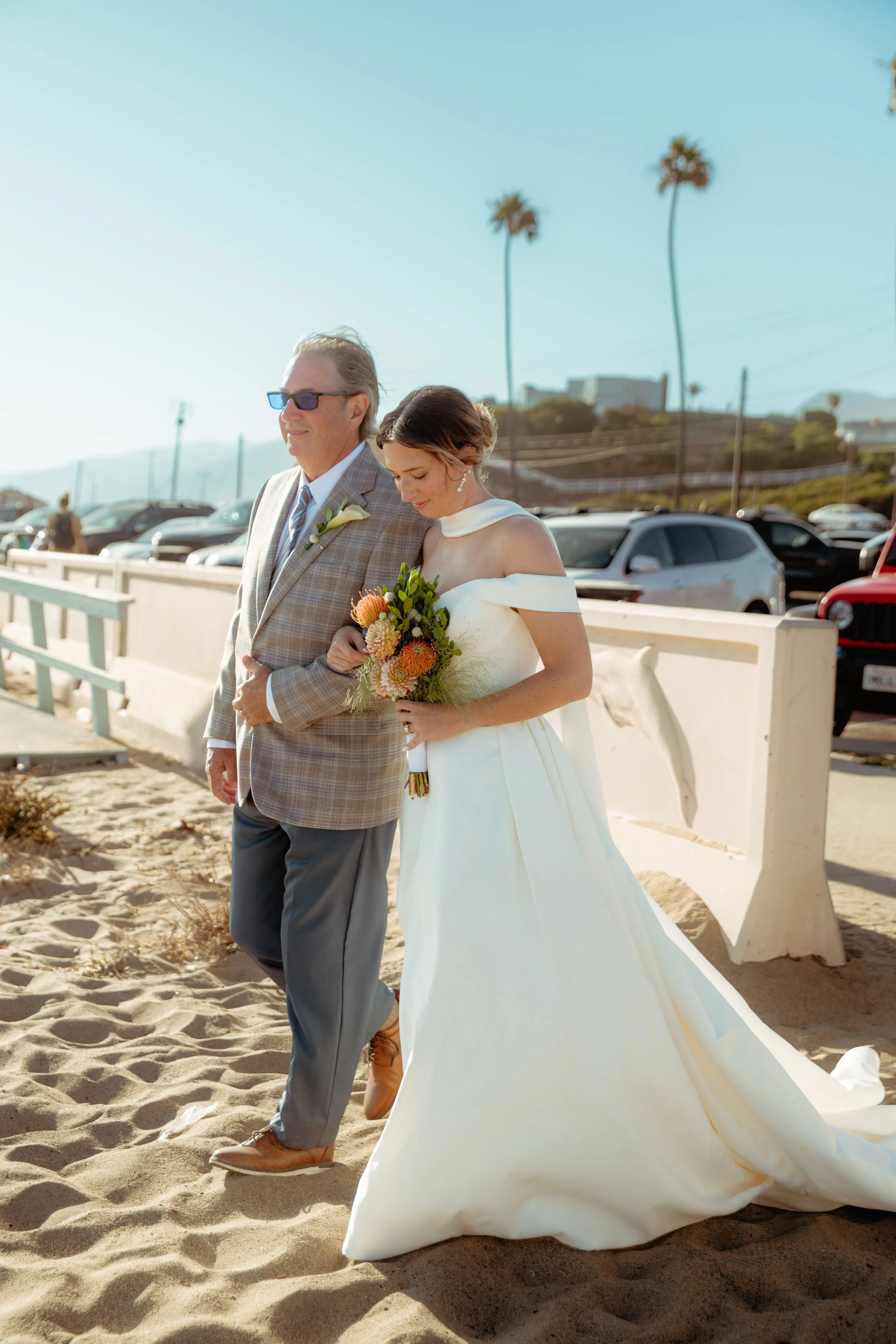 Bride being walked by her father onto the sand at Zuma Beach with palm trees behind her.