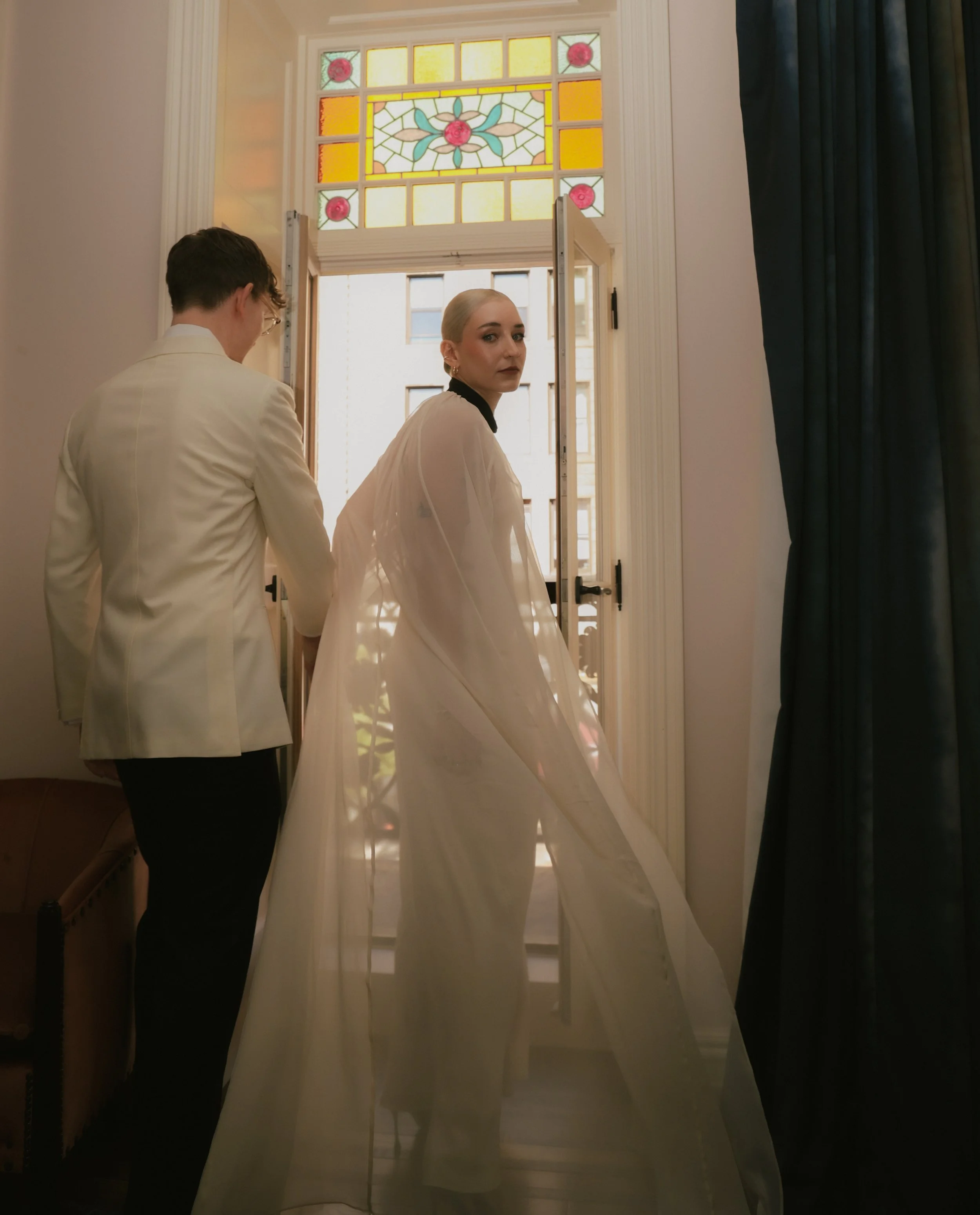 Couple looks back in front of stained glass window inside of the Chelsea Hotel whre they pose for editorial style photography at the Chelsea Hotel