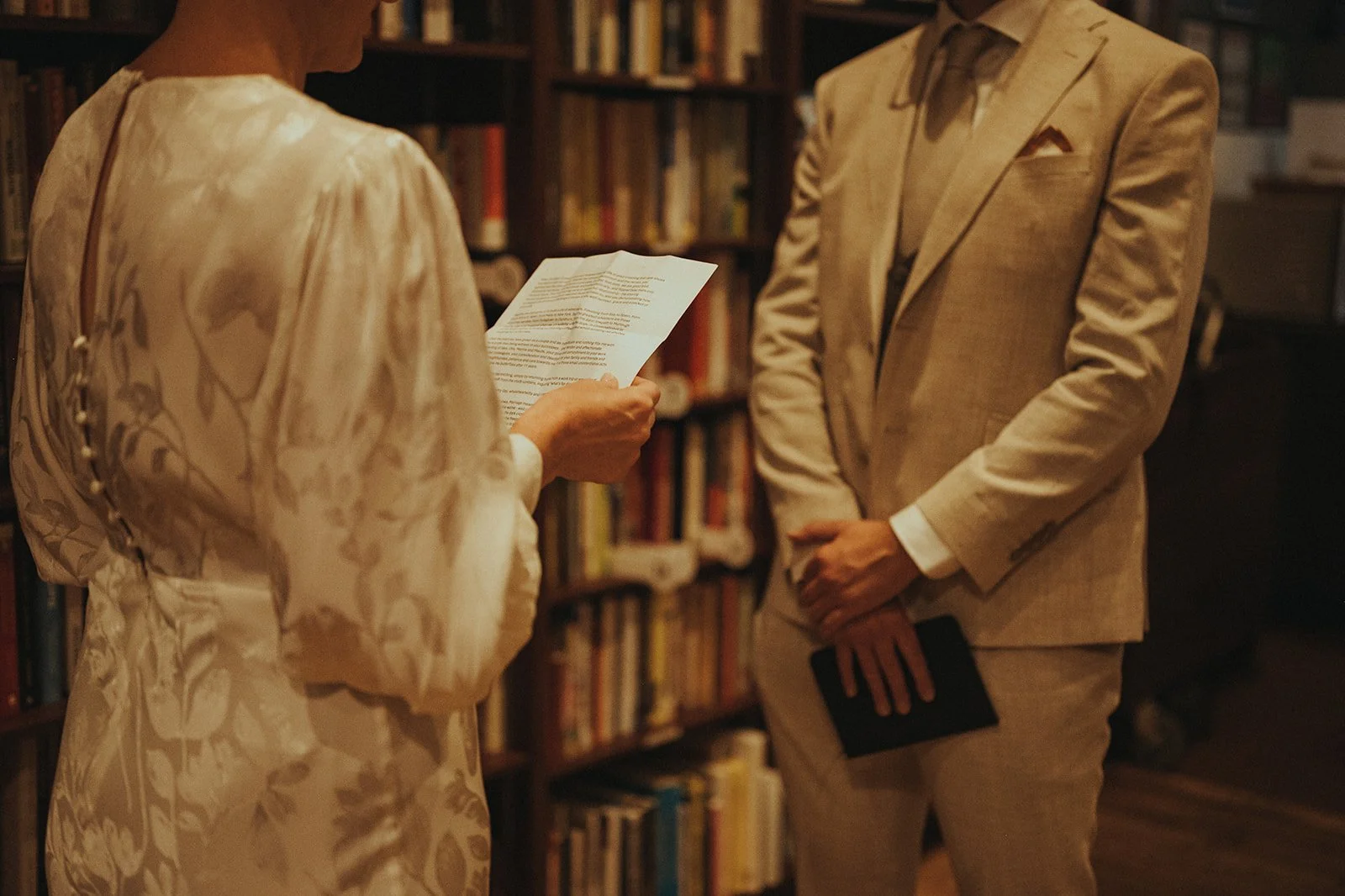 Bride and groom reading each other vows in front of tall bookcases inside Housing Works Bookstore in SoHo during Cakewalk elopement ceremony.