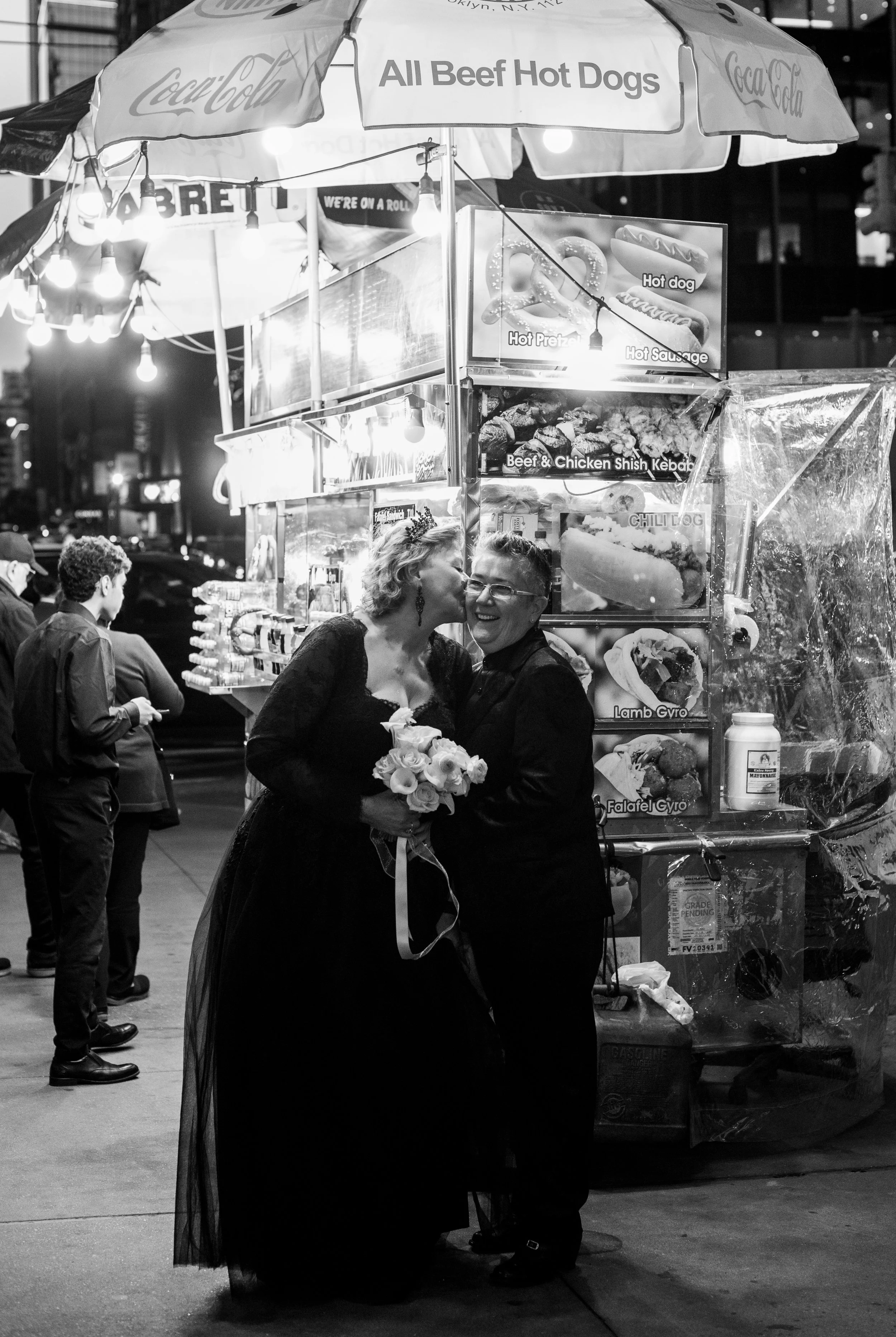 Two women wearing black and holding a yellow bouquet embracing under a Manhattan hot dog stand after their LGBTQ Cakewalk elopement.