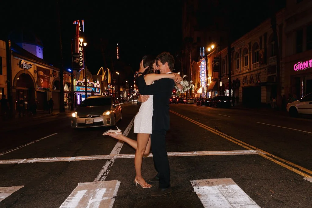 Couple crossing the street on Hollywood Blvd at night in wedding attire with the marquis lights in the distance.