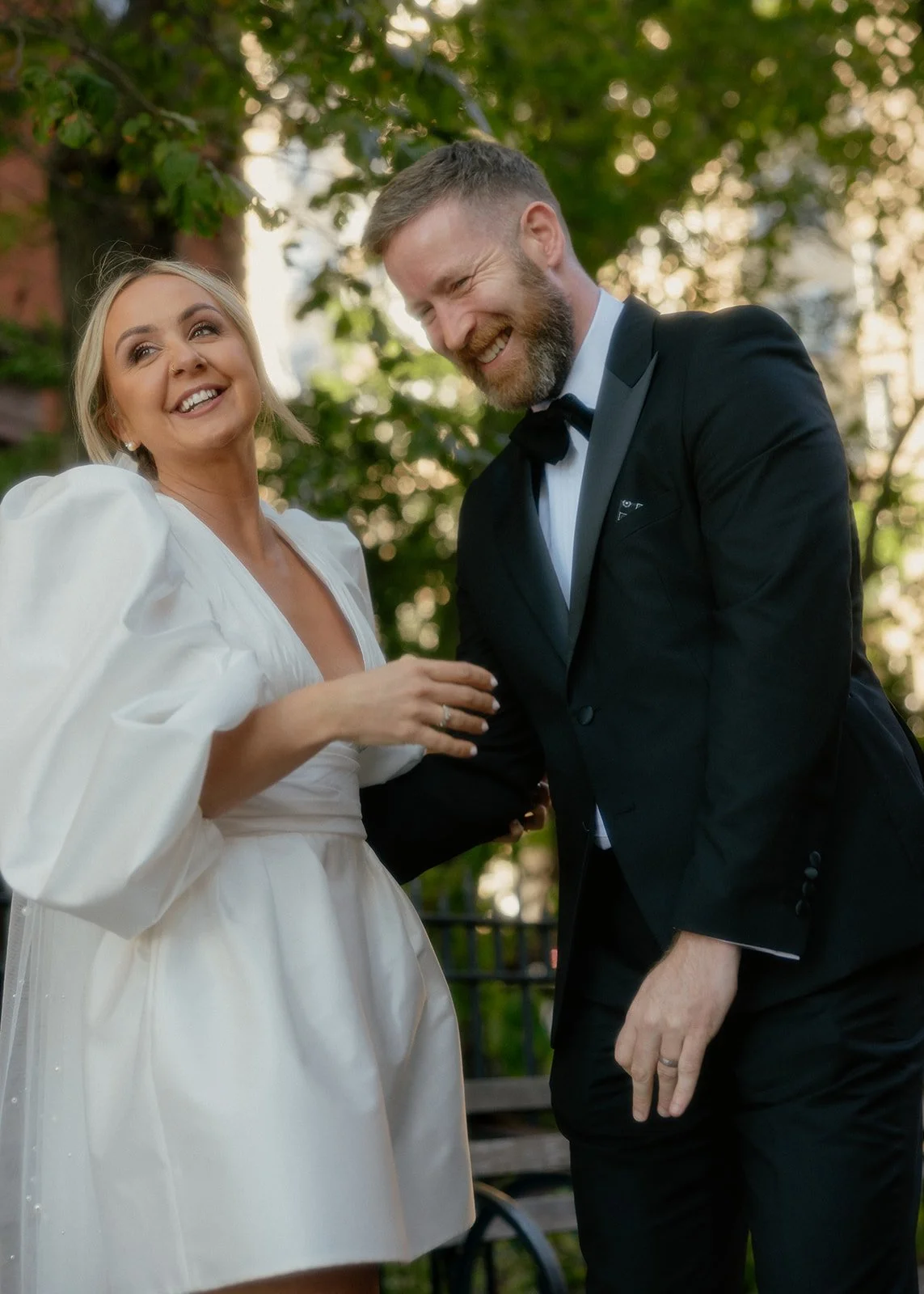 A smiling woman in a white wedding dress and a man in a black tuxedo are standing outdoors, laughing and holding hands, in a park setting with trees and sunlight in the background.