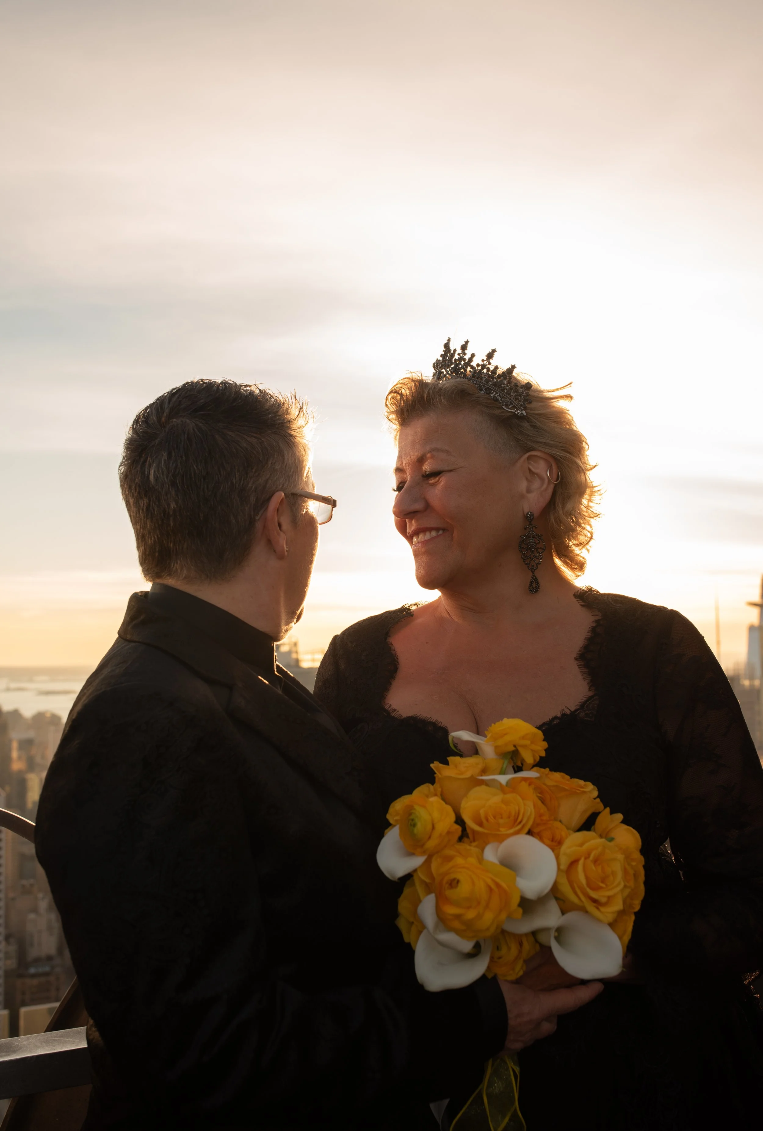 Two women wearing black and holding a yellow bouquet face each other smiling at the Top of the Rock before their LGBTQ Cakewalk elopement.