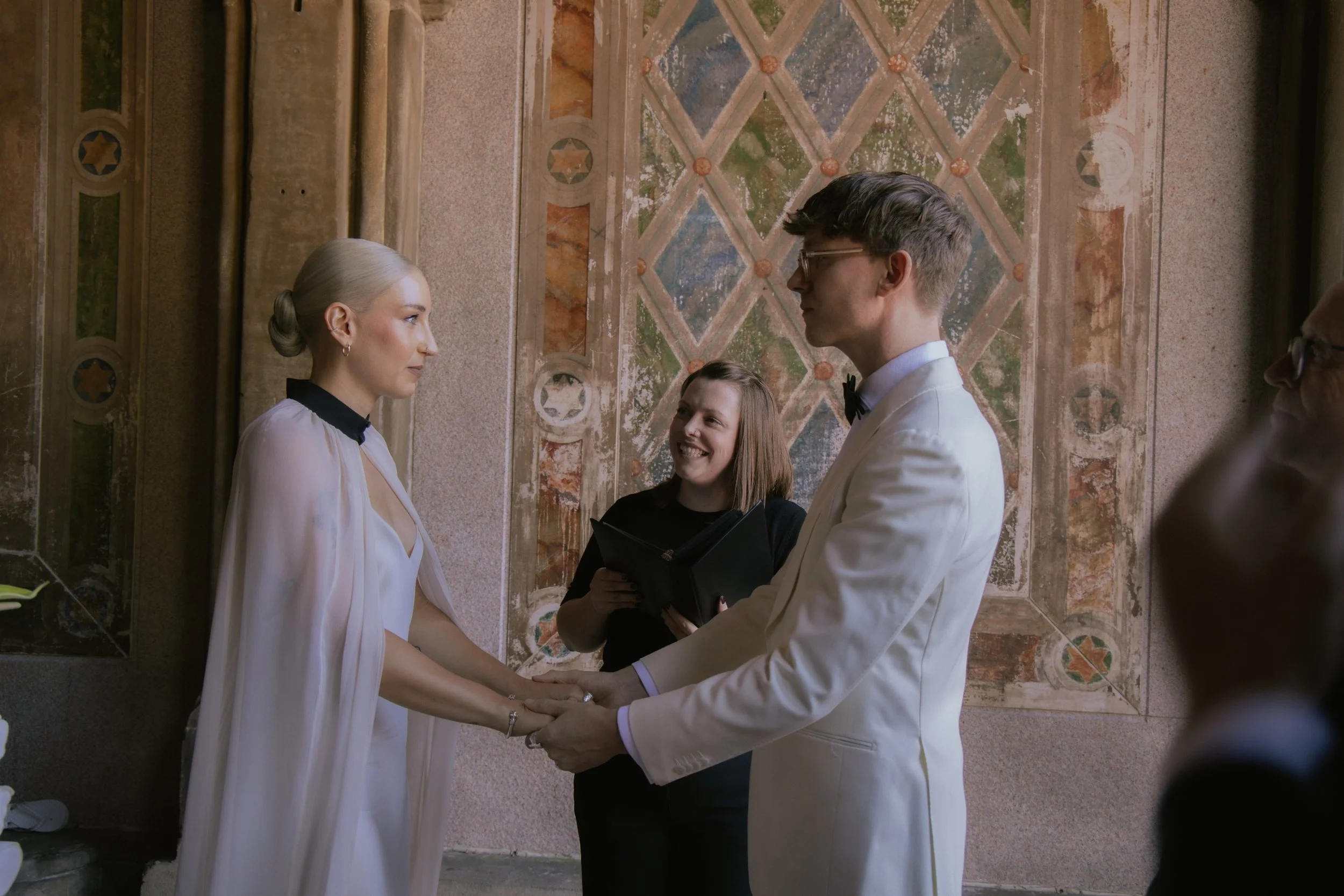 Bride and groom hold hands while officiant delivers ceremony speech at Bethesda Terrace in Central Park at Cakewalk elopement.
