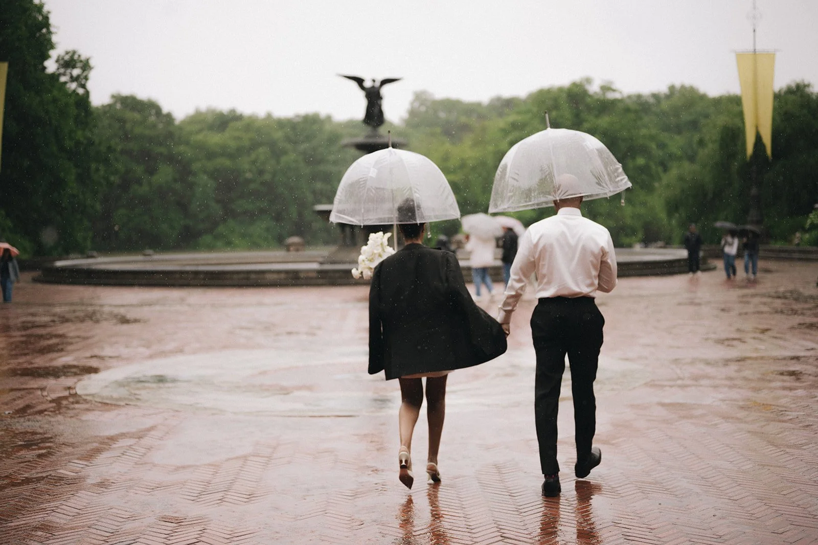 Bride and groom holding clear umbrellas, walking toward the fountain at Bethesda Terrace after their Cakewalk elopement ceremony.