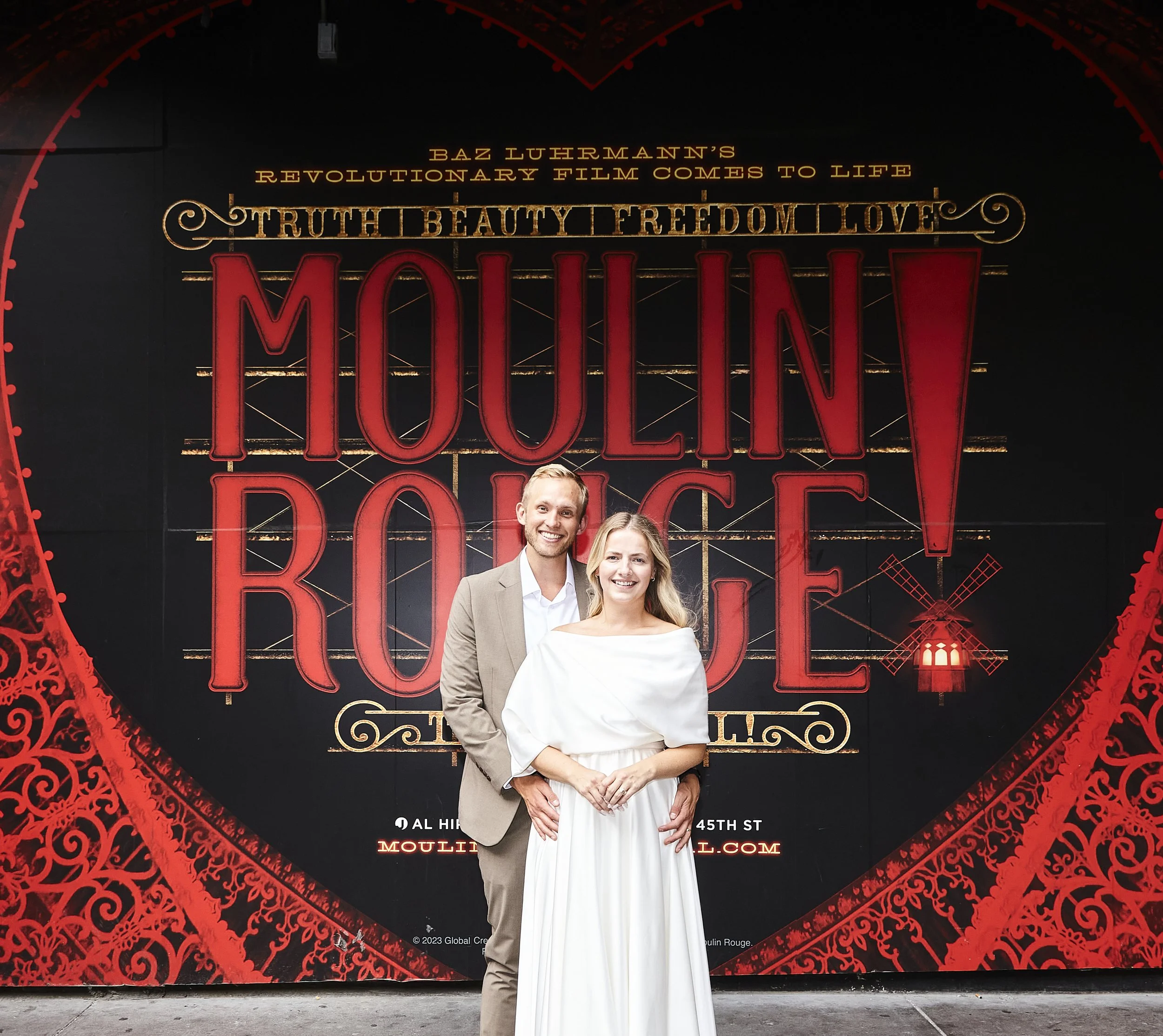 Bride and groom posing in front of a Moulin Rouge! on Broadway poster after their Cakewalk elopement.