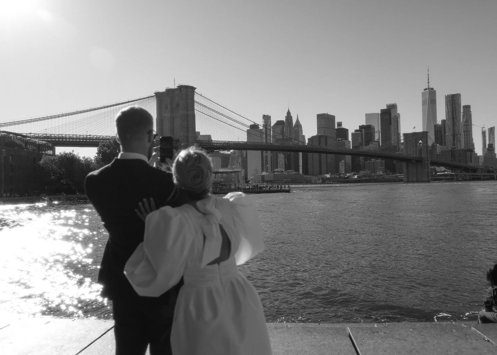 Bride and groom admiring the Brooklyn Bridge and the Manhattan skyline from the edge of the water.