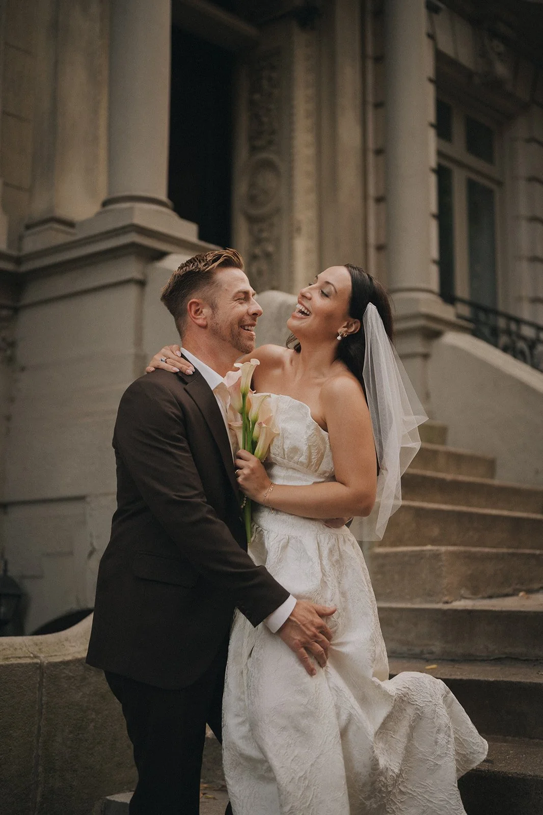 A happy couple on their wedding day, standing on stairs outside a historic building, with the bride in a white dress holding calla lilies and the groom in a suit, sharing a joyful moment.