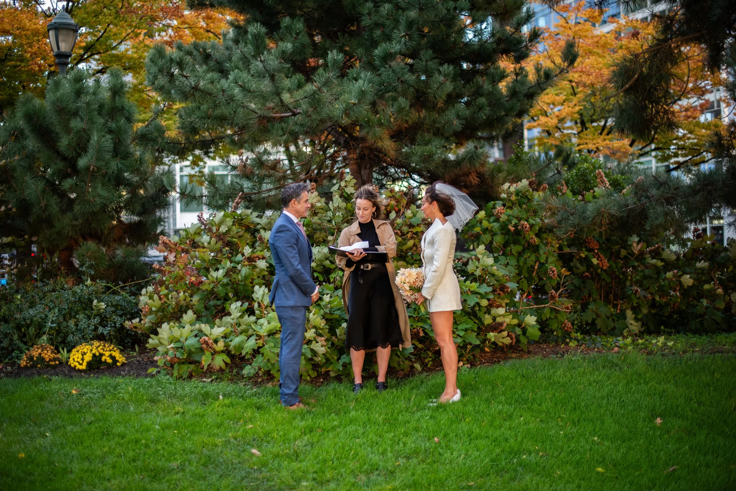 Bride and groom being married at Hudson River Park on the grass, bouquet in hands, with Cakewalk officiant reading speech behind them.