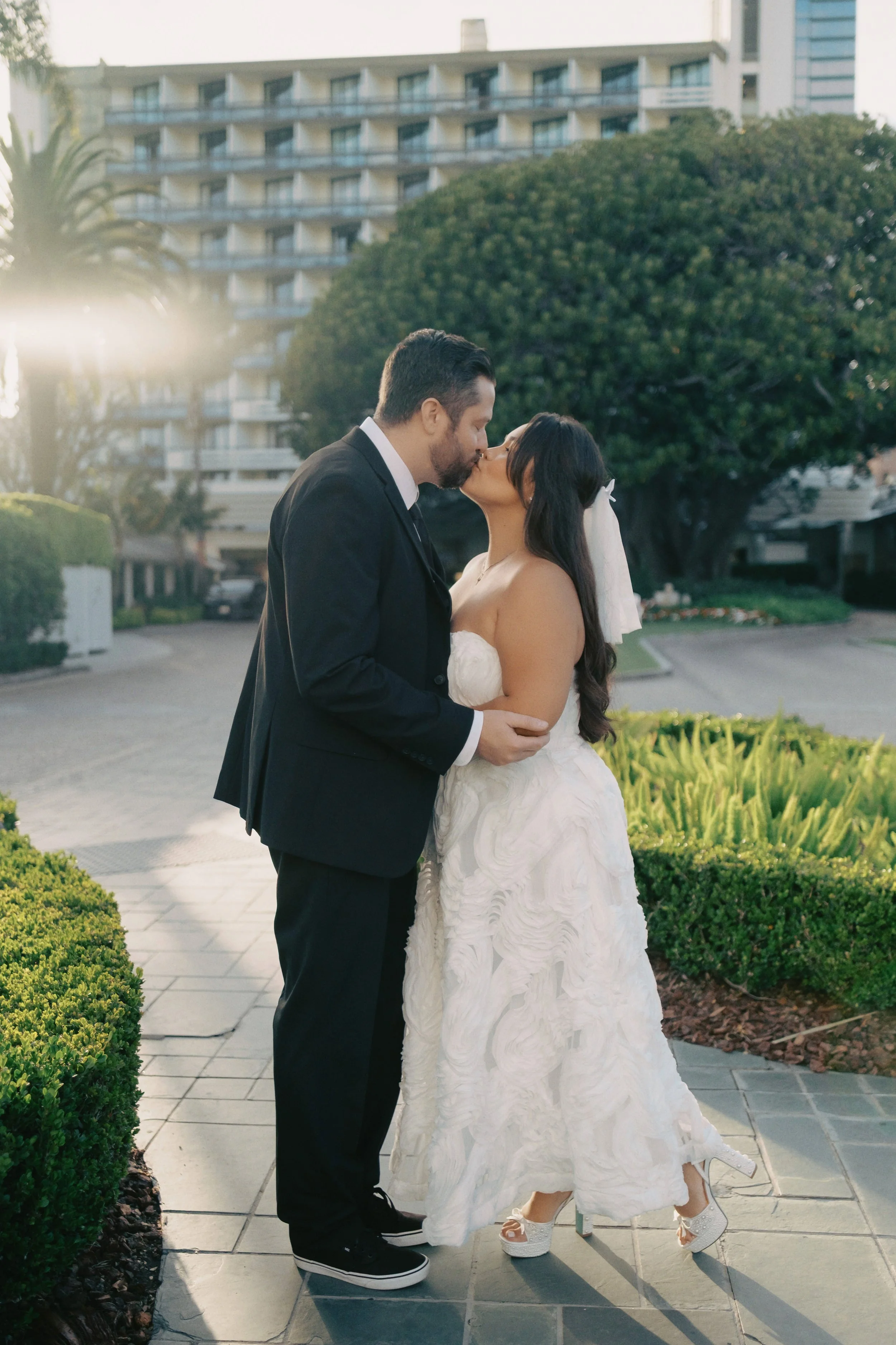 Bride and groom kissing in front of a tree on a paved path at the Fairmont Miramar in Santa Monica before their Cakewalk elopement ceremony.