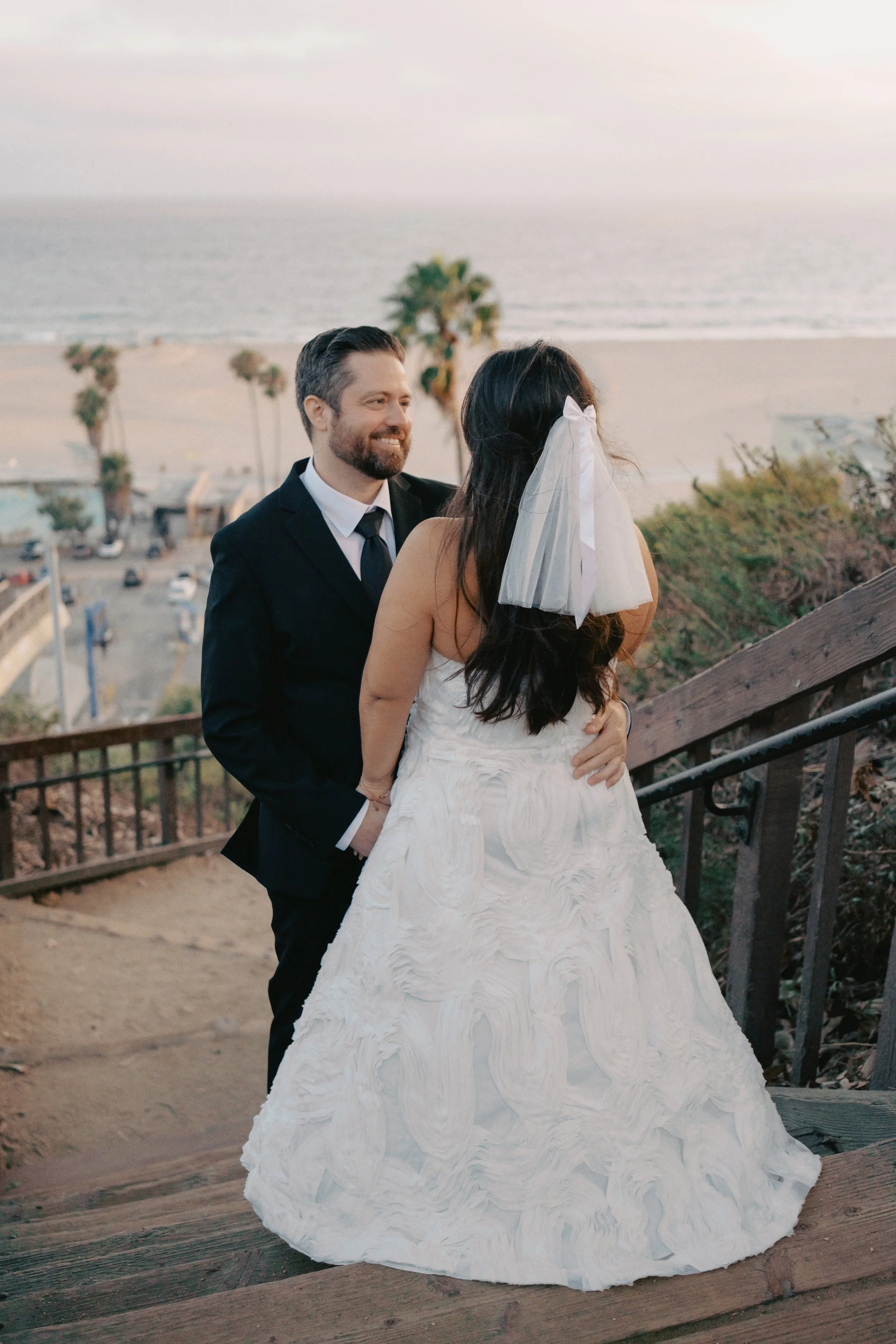 Bride and groom standing on cliffside stairs in Santa Monica with the palms trees, the sand, and the ocean behind them.