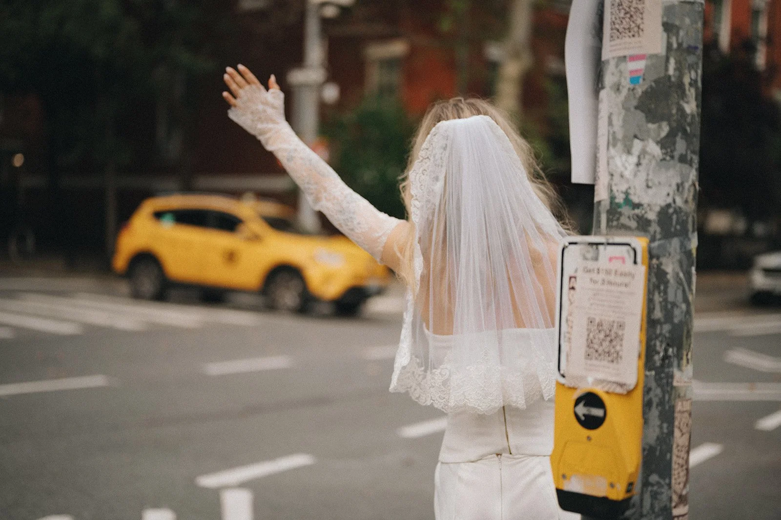 A person dressed in a wedding gown with a veil, standing on a city street at night, waving with one hand and standing near a utility pole with a bus stop sign.