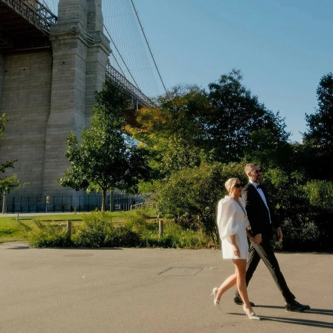 A man and woman dressed formally walking outdoors near the Brooklyn Bridge in New York City, with trees and greenery in the background.