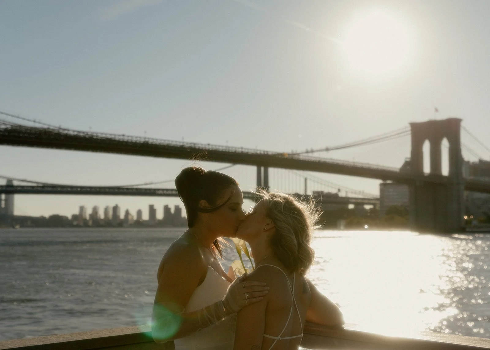 LGBT brides kisses with the Brooklyn Bridge in the distance as the sun rises and shines over the river after their Cakewalk elopement.