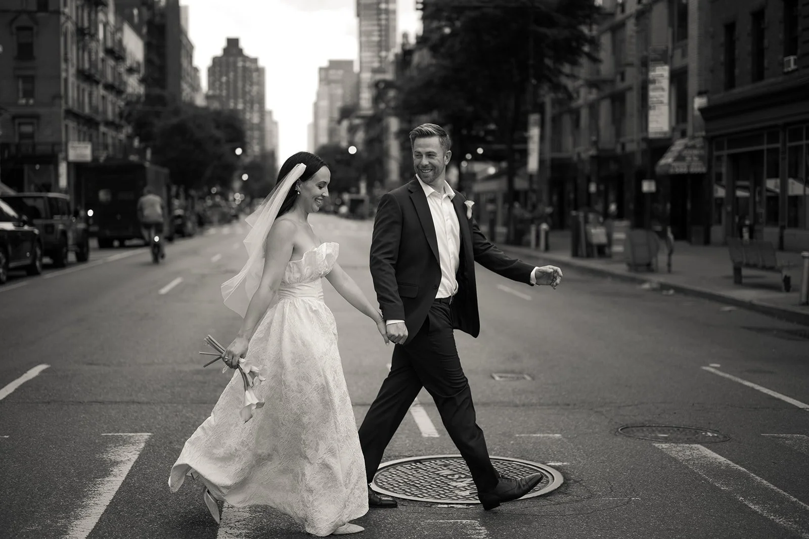 A black and white photo of a bride and groom walking hand in hand in the middle of a city street, smiling and joyful.