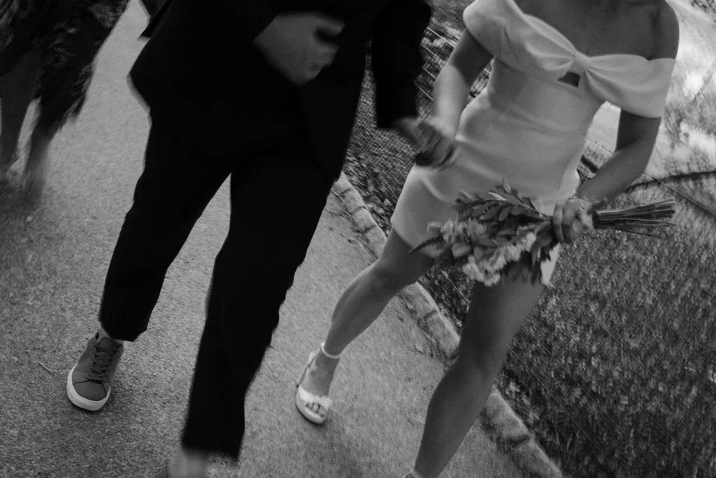 A woman in a white dress holding a bouquet of flowers, standing next to a man in a dark suit. The setting appears to be outdoors, possibly a wedding or special event.