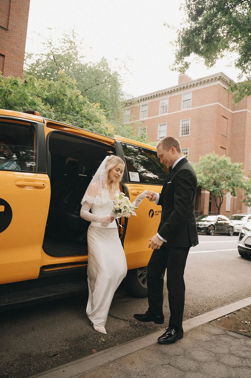 A bride in a white dress and veil holding a bouquet, and a groom in a black tuxedo exchanging a ring in front of a yellow taxi on a city street with trees and brick buildings in the background.