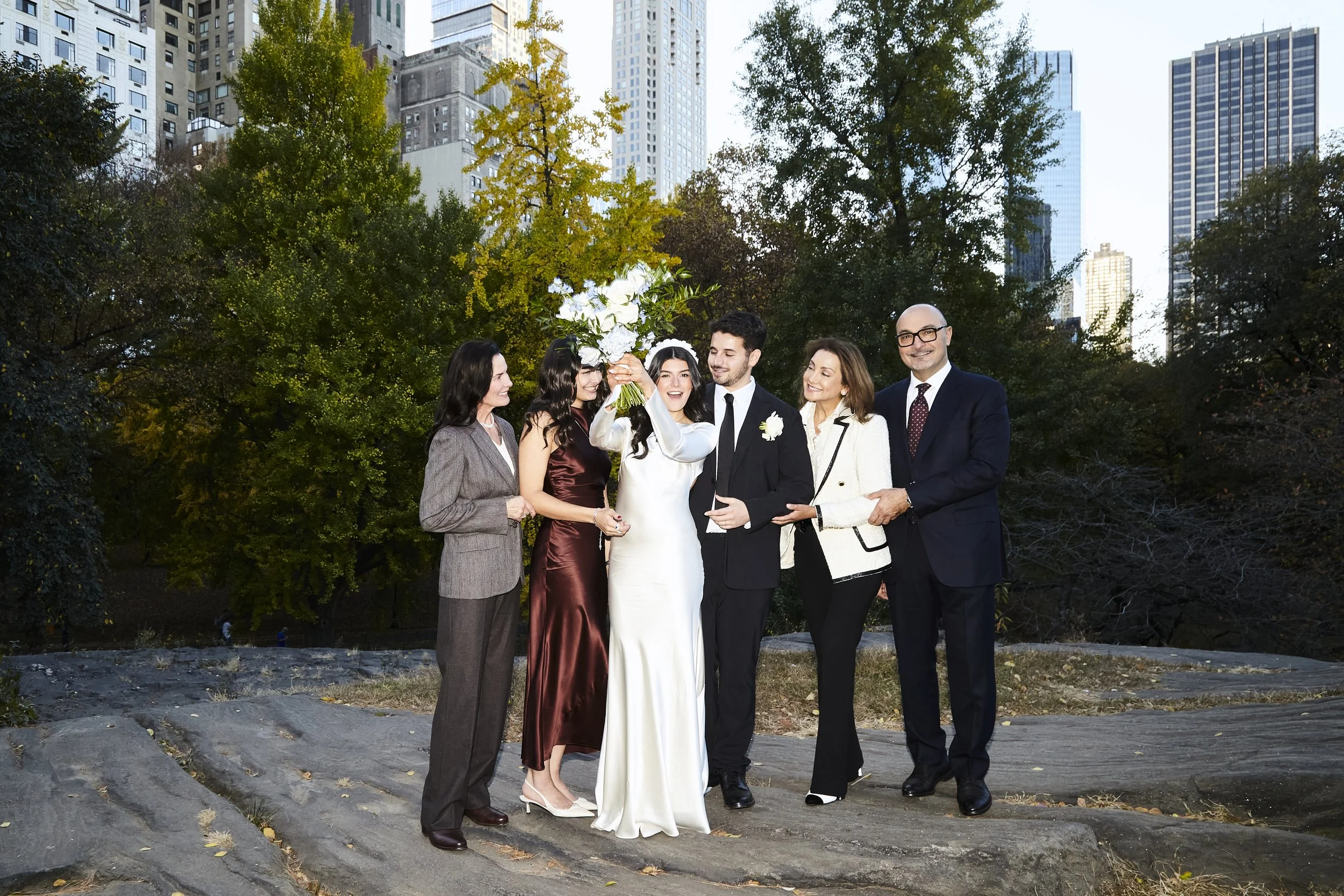 Bride, groom and family standing in Central Park with fall foliage and the city skyline behind them after their Cakewalk micro wedding.