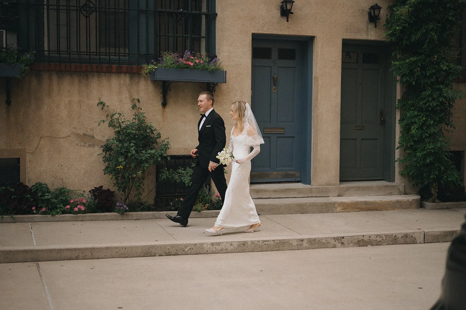 A newlywed couple walking on the sidewalk outside a building. The groom is wearing a black tuxedo and the bride is wearing a white wedding dress with lace sleeves, holding a bouquet of flowers. The building has blue doors, a planter with pink and pur