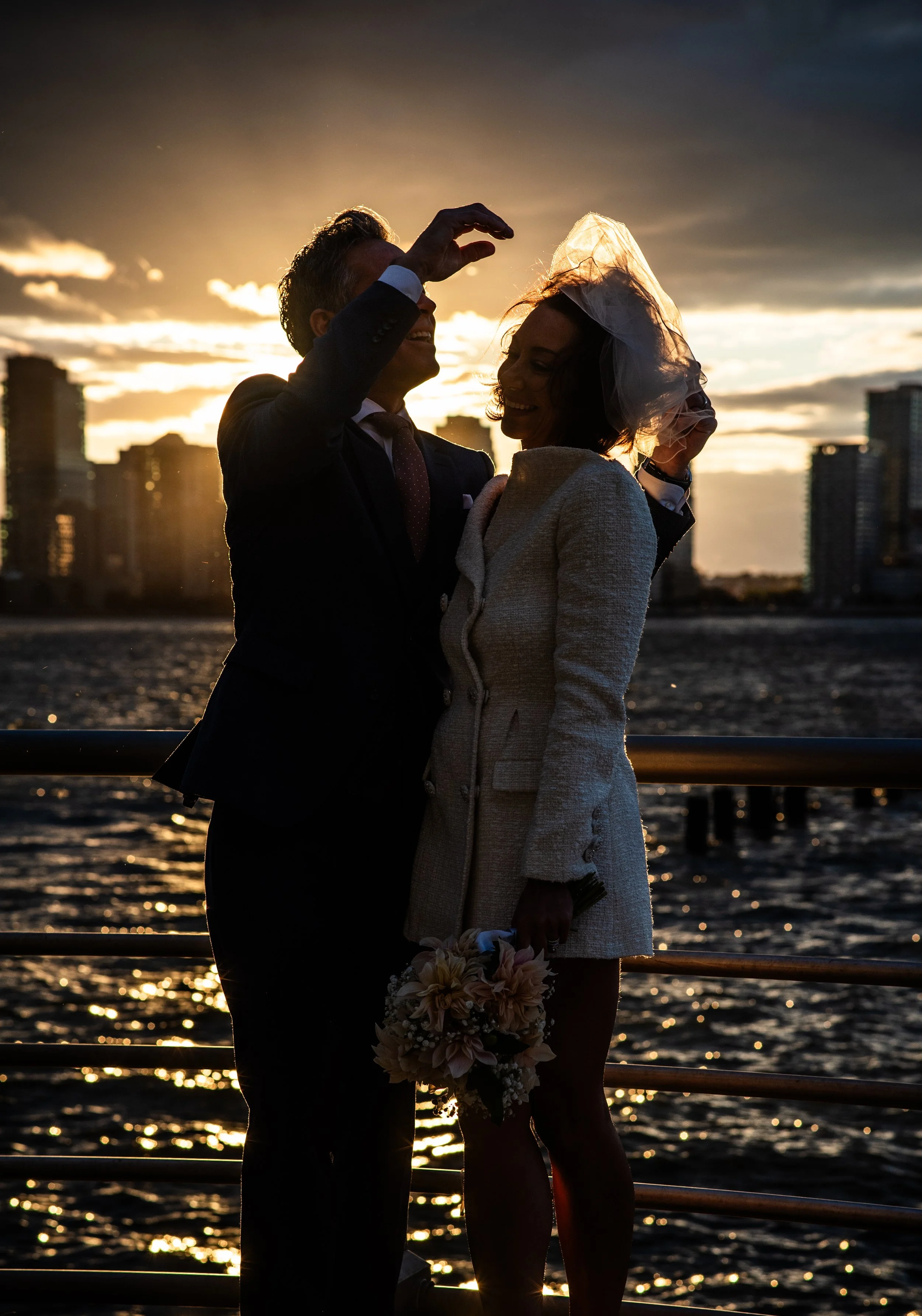 Groom helping bride with her veil as the sun sets at Hudson River park, the glow of the day behind them, reflecting off the water.