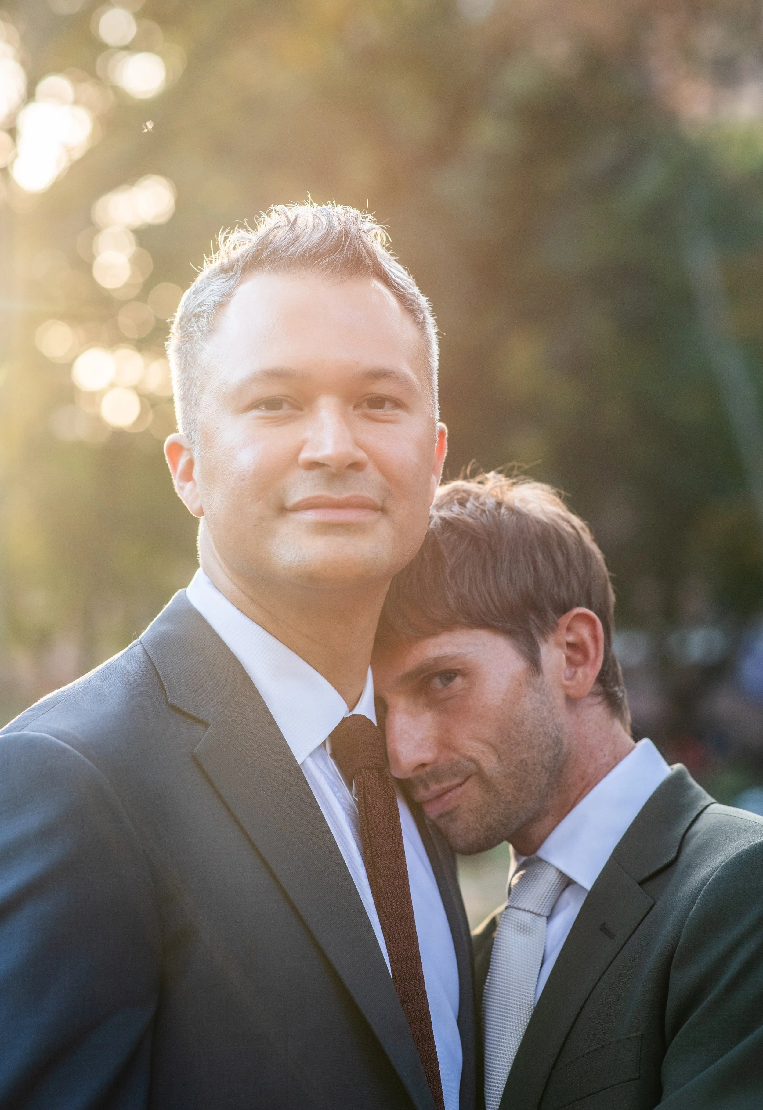 Two men in Washington Square Park embracing with the trees and sunlight behind them.