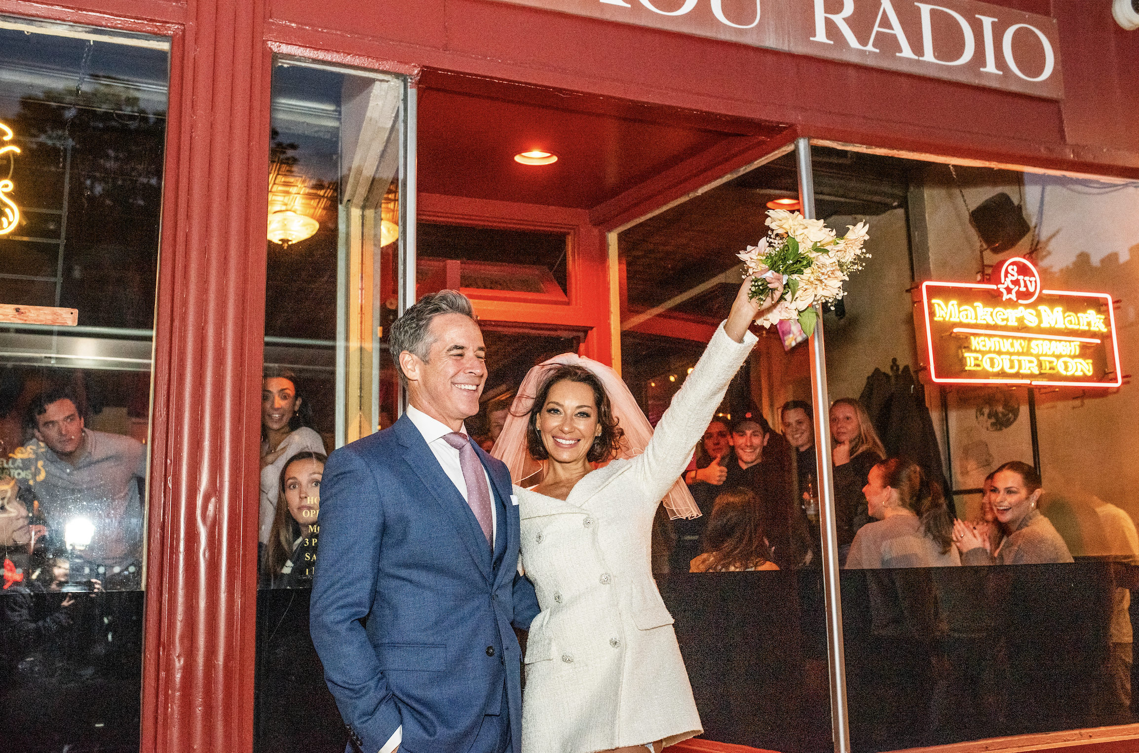 A bride in a white coat holding a bouquet raises her arm in celebration, standing next to a smiling groom in a blue suit outside a restaurant with a neon sign reading 'Maker's Mark Kentucky Straight Bourbon,' while a group of friends inside cheers.