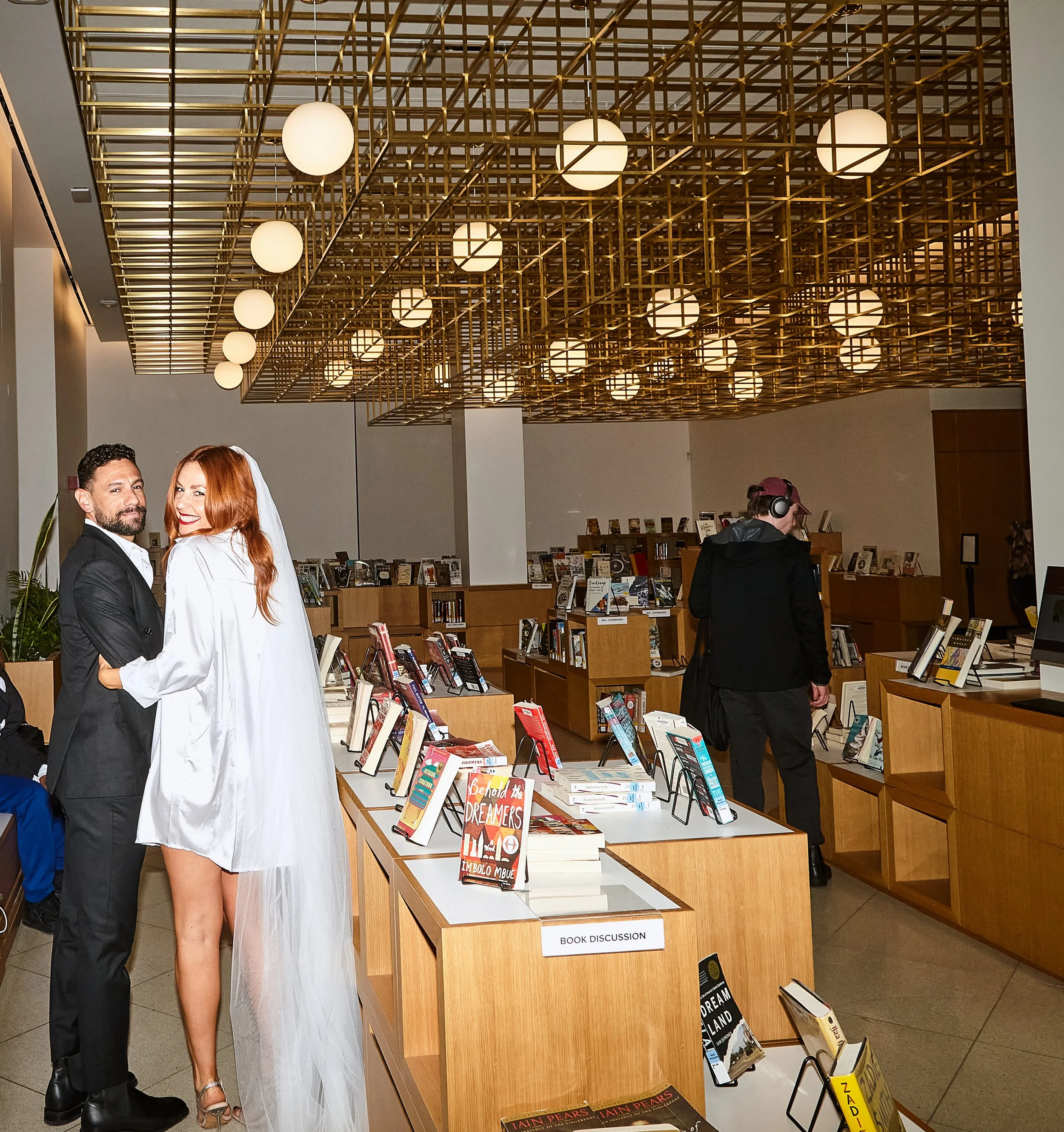 Bride and groom embracing inside Brooklyn Central Library after their Cakewalk ceremony. Lights are on the ceiling and book displays are spread about.