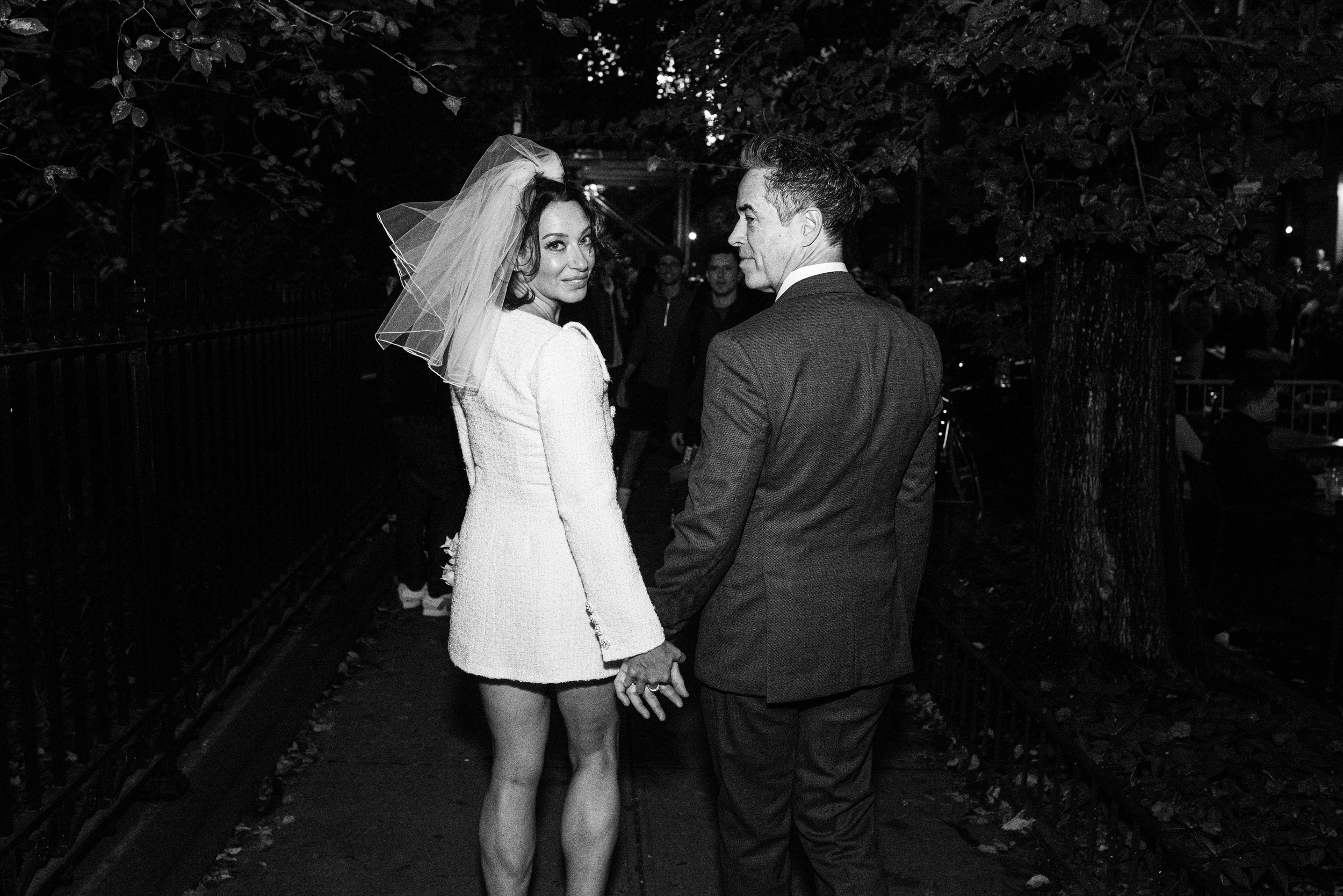 A woman in a wedding dress and veil holding hands with a man in a suit, walking outdoors at night, with trees and people in the background.