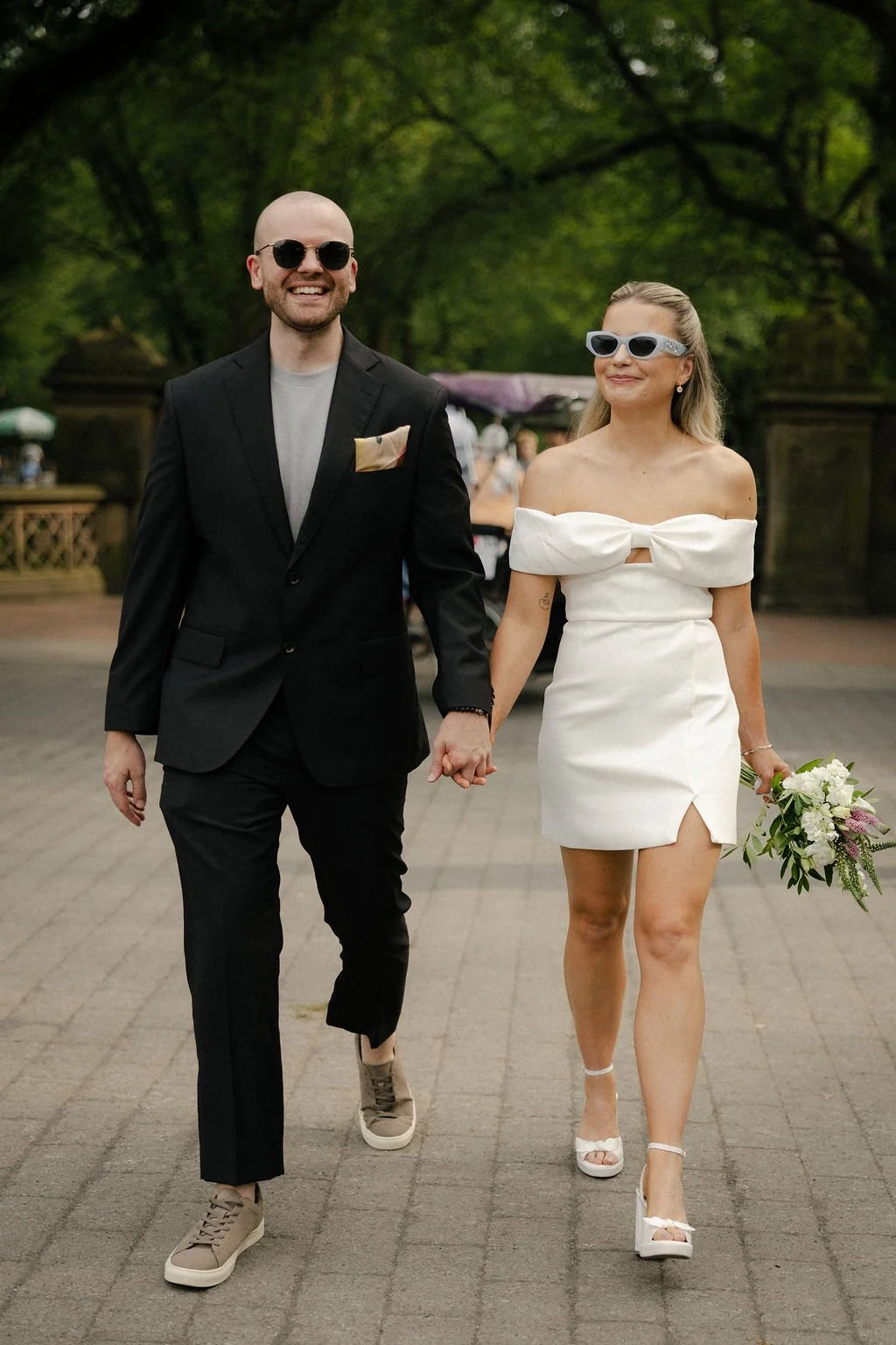 Bride and groom walking on pavement to in Central Park with greenery behind them, on the way to their Cakewalk elopement.
