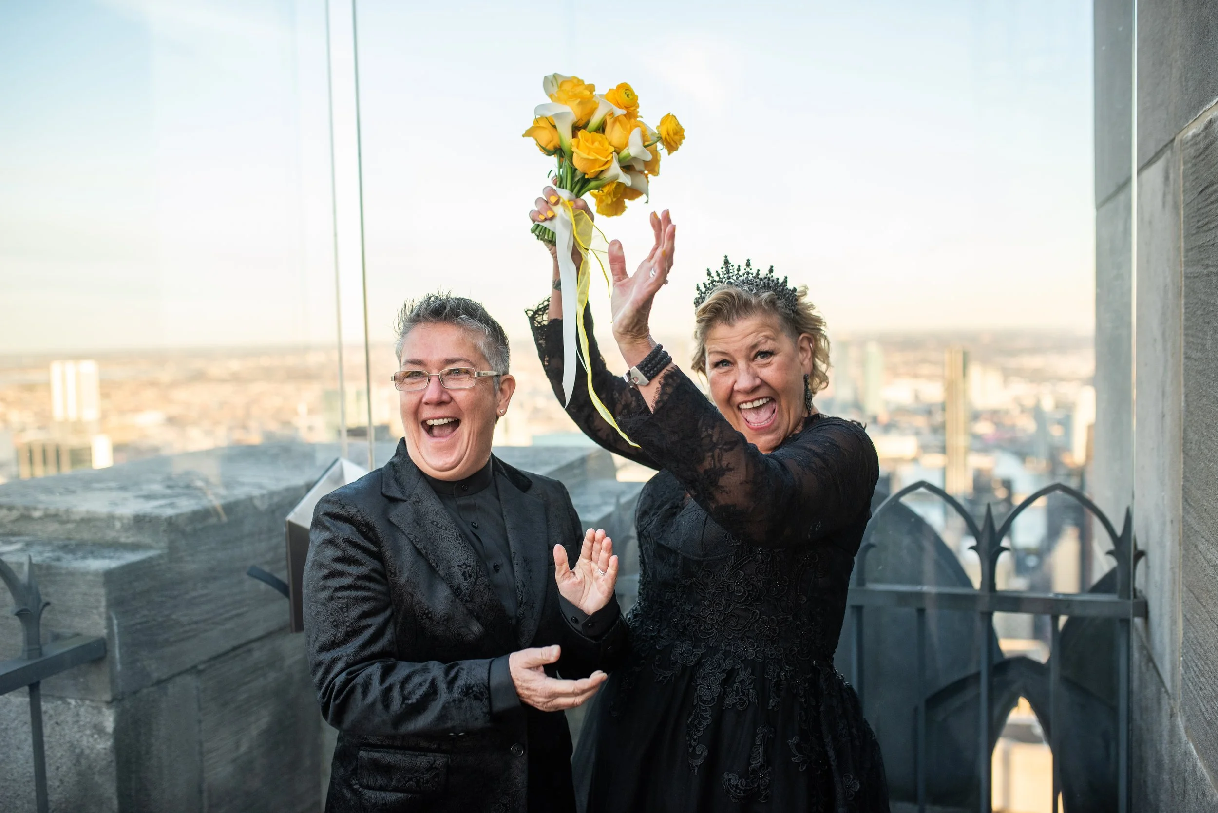 Two women wearing black and holding a yellow bouquet clapping and cheering at the Top of the Rock after their Cakewalk elopement ceremony.
