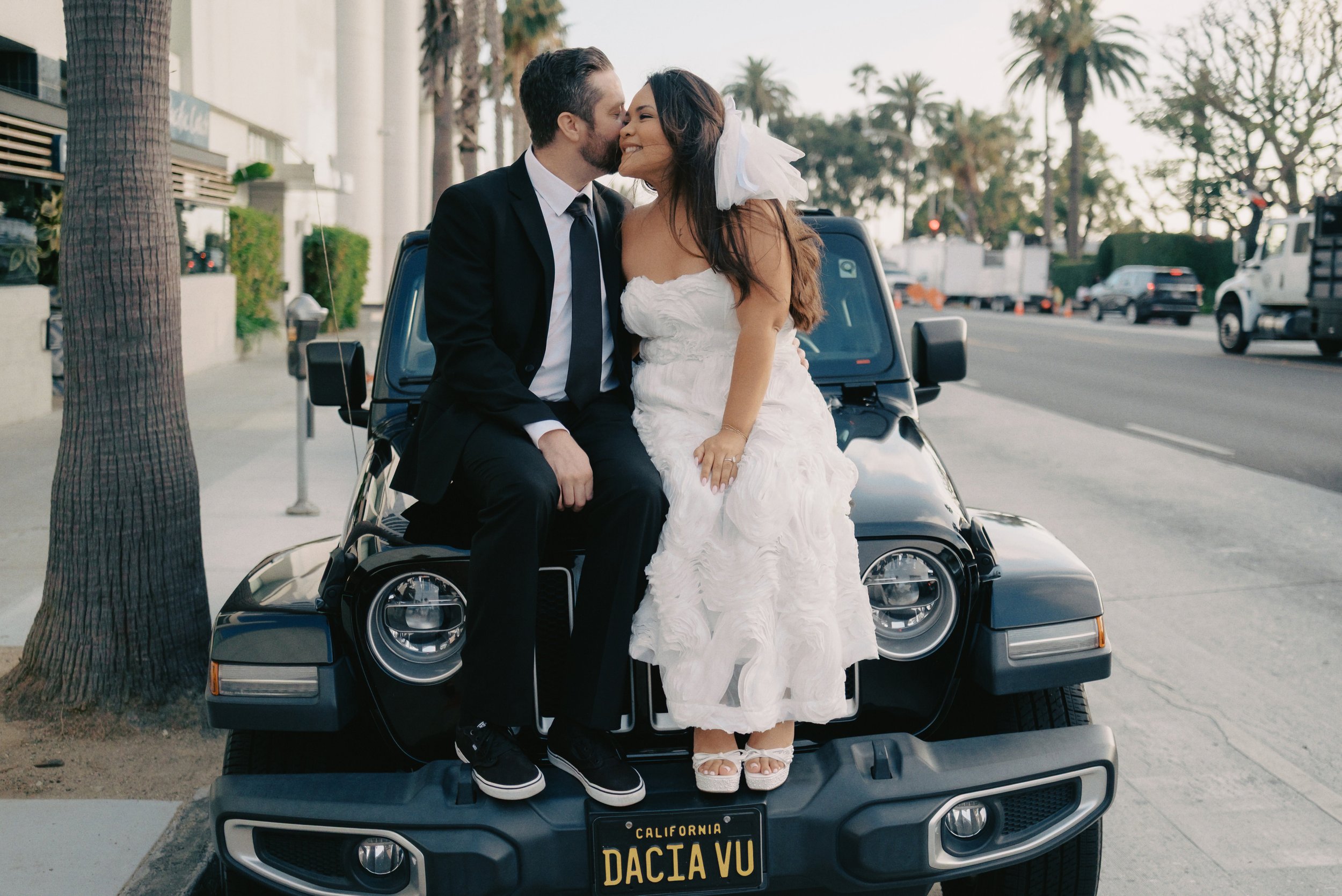 A newlywed couple sitting on the hood of a black car with a California license plate reading 'Dacia Vu.' The groom is kissing the bride on the cheek, and she is smiling, wearing a white dress with a veil. They are outdoors on a city street with palm trees and parked vehicles in the background.