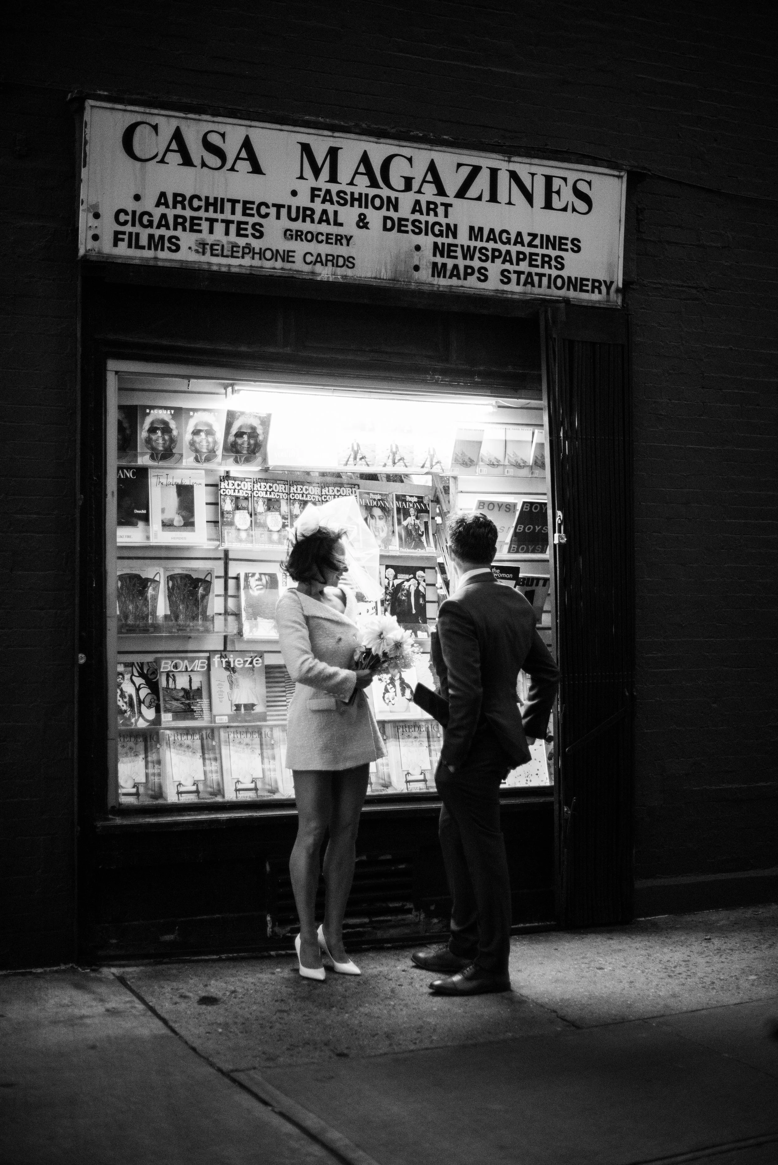 A woman in a coat and high heels holding a bouquet of flowers talking to a man in a suit outside a magazine store at night.