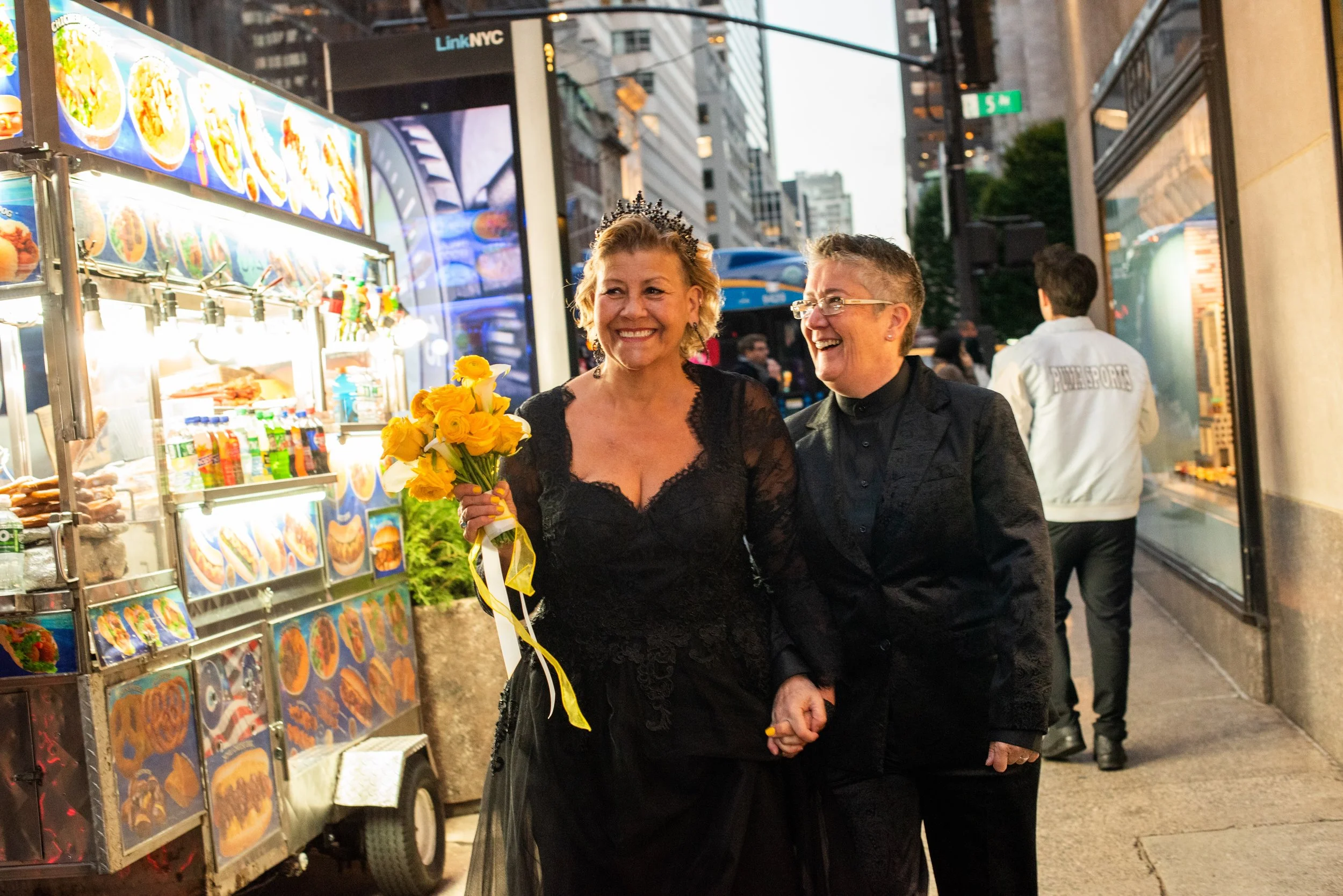 Two women wearing black and holding a yellow bouquet smiling as they walk down the Manhattan street near a food truck after their LGBTQ Cakewalk elopement.