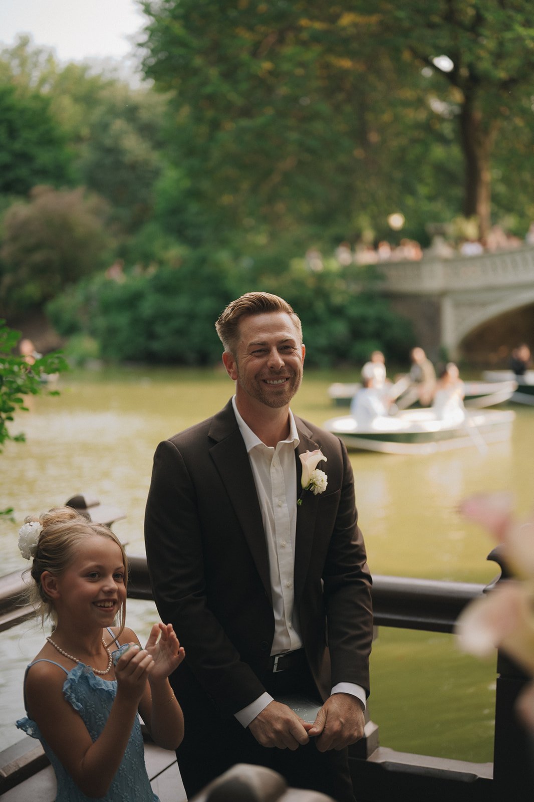 Groom and daughter stand in front of the water at Bow Bridge waiting for bride at Central Park elopement ceremony.