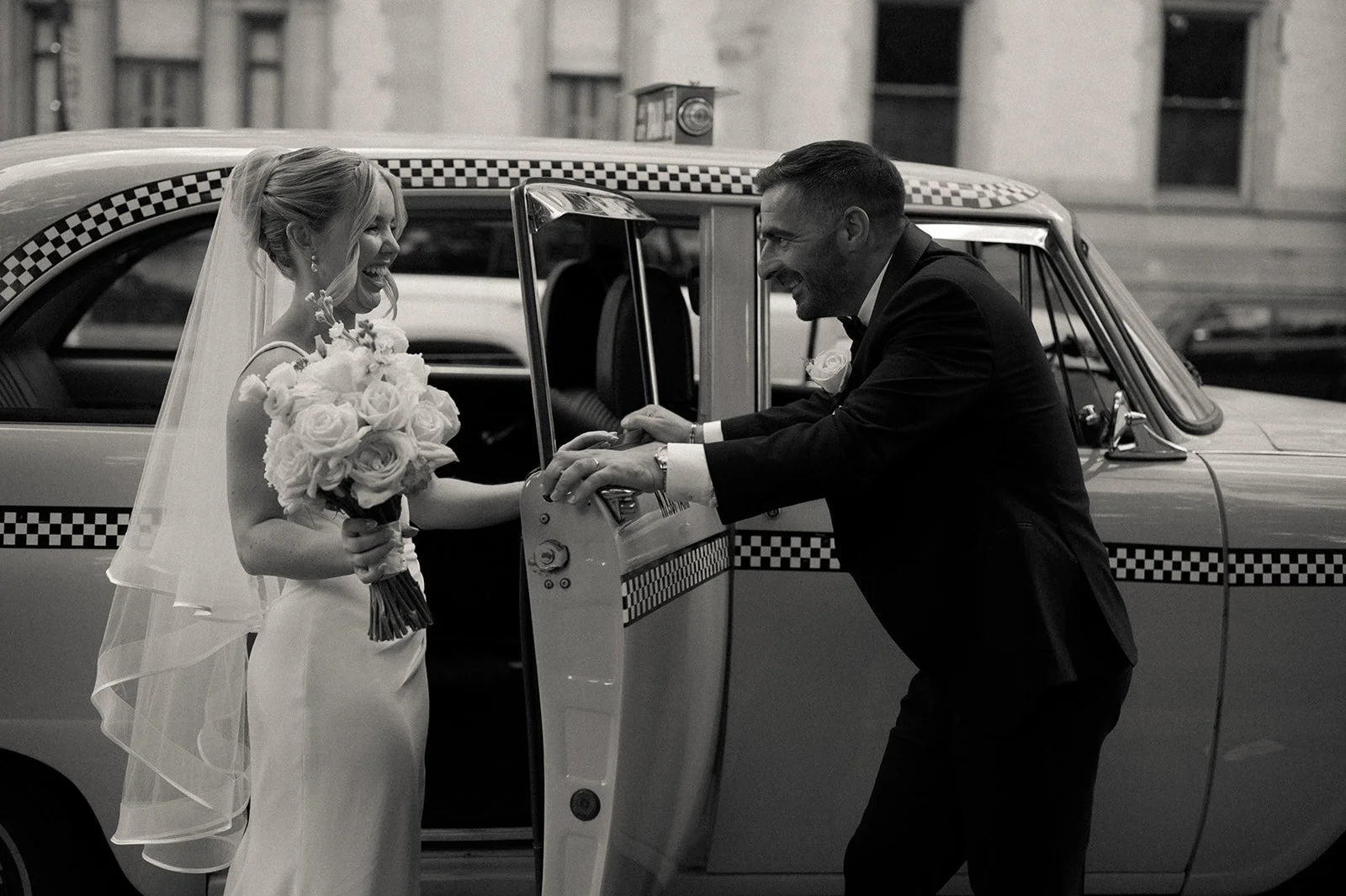 Bride and groom looking at each other through a vintage checkered taxi cab window, smiling and holding a white bouquet.