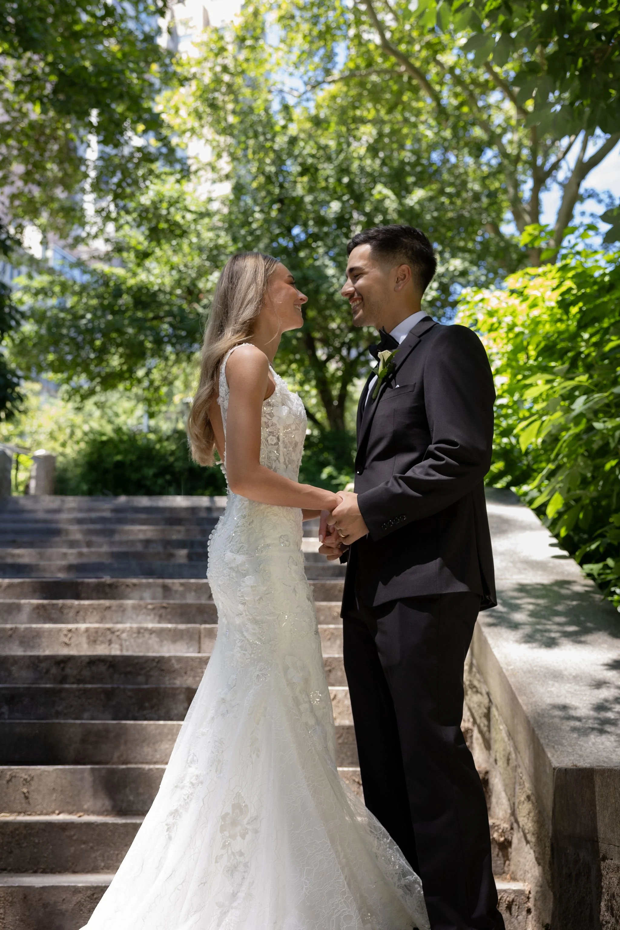 Bride and groom standing on stairs in Central Park looking at each other, surrounded by greenery.