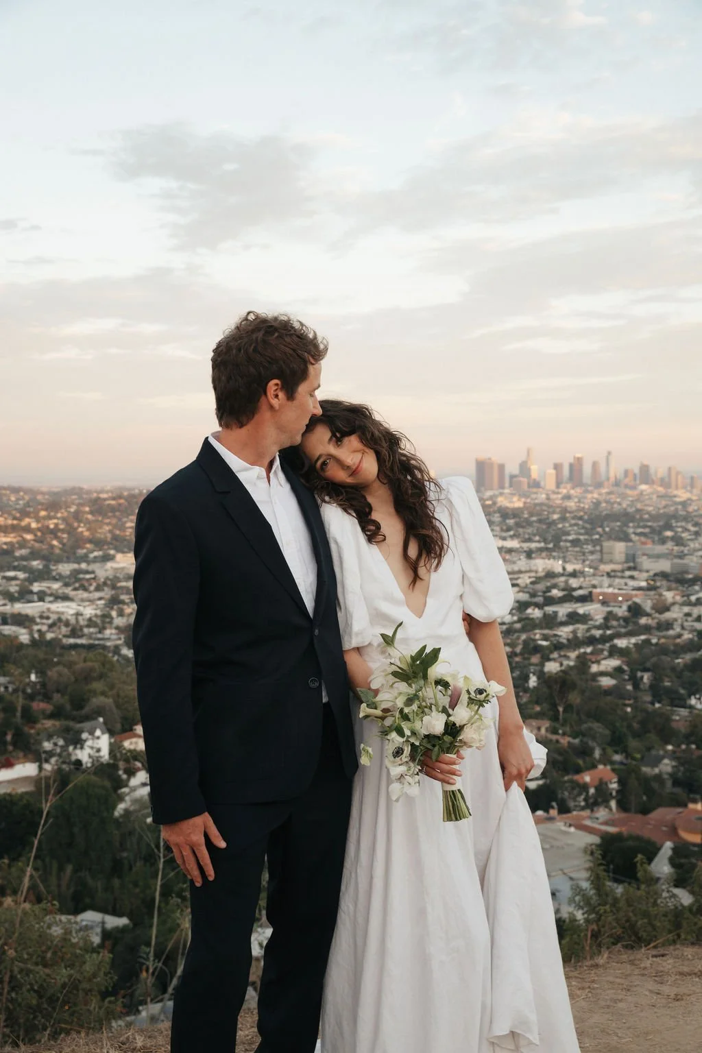 A couple dressed in wedding attire standing closely together on a hilltop overlooking a cityscape at sunset. The woman is holding a bouquet of white flowers and leaning her head on the man's shoulder.