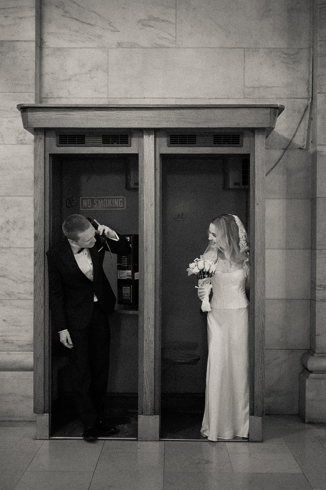 A black and white photo of a bride in a wedding dress and veil holding a bouquet of roses and a groom in a tuxedo chatting inside a small wooden phone booth with a 'No Smoking' sign on the wall behind them.