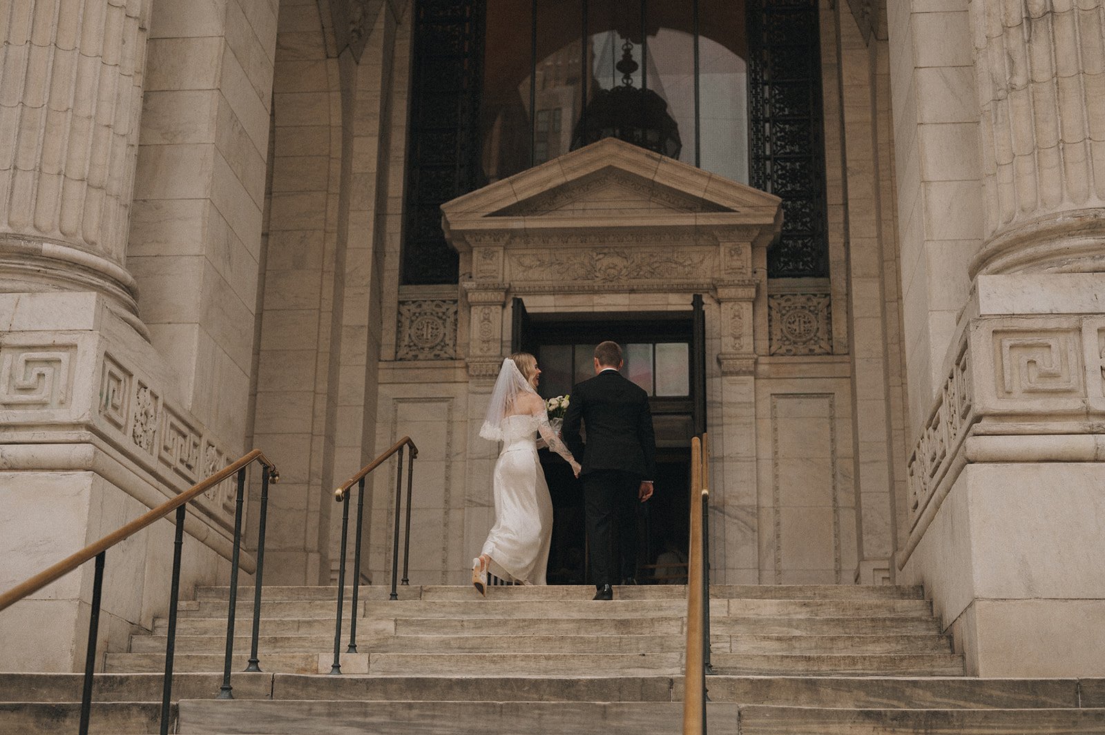A bride and groom walking up the steps of a grand stone building, holding hands. The bride is wearing a white wedding dress with a veil, and the groom is dressed in a black suit. The building features large stone columns and intricate stonework.