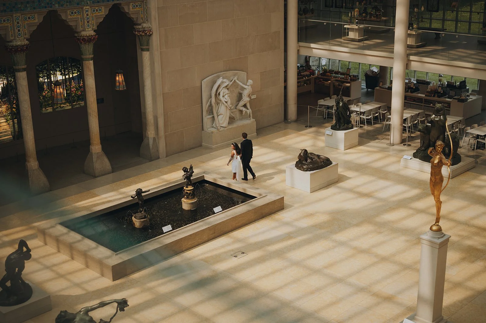 Bride and groom walking inside the Met Museum of Art among the sculptures and a fountain.