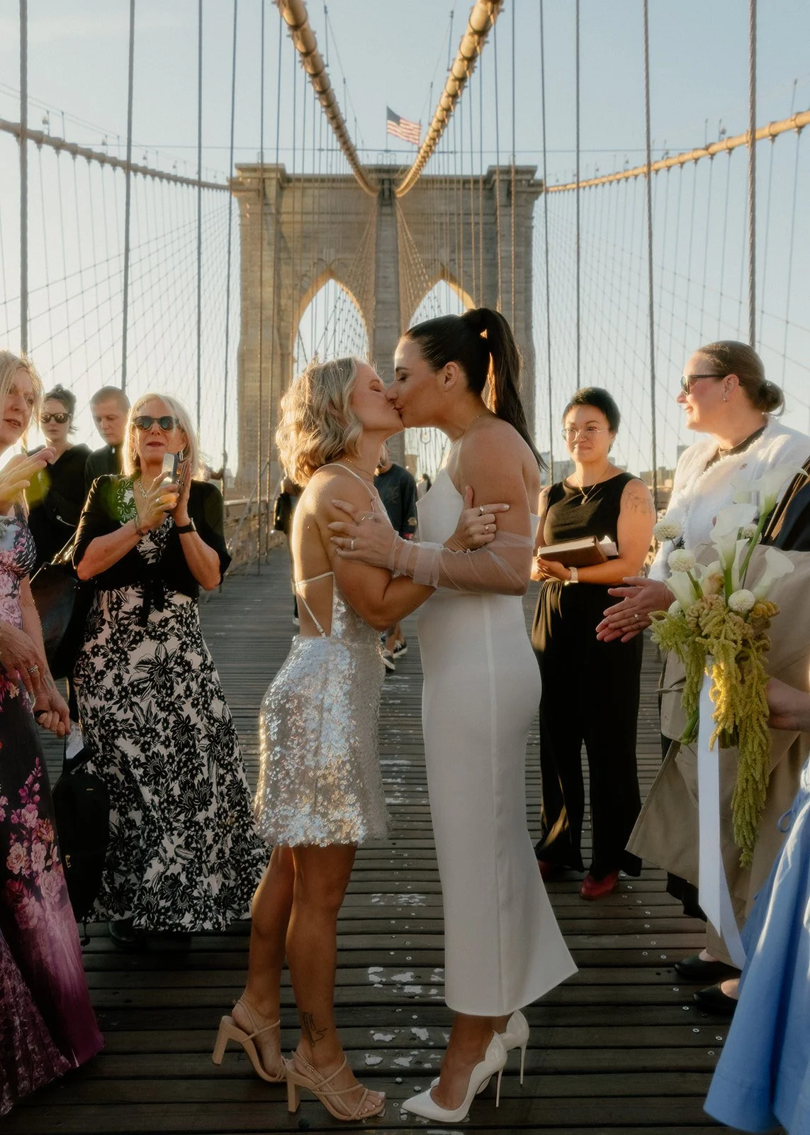 LGBT brides kissing at their Cakewalk elopement on the Brooklyn Bridge at sunrise surrounded by their group of guests cheering.