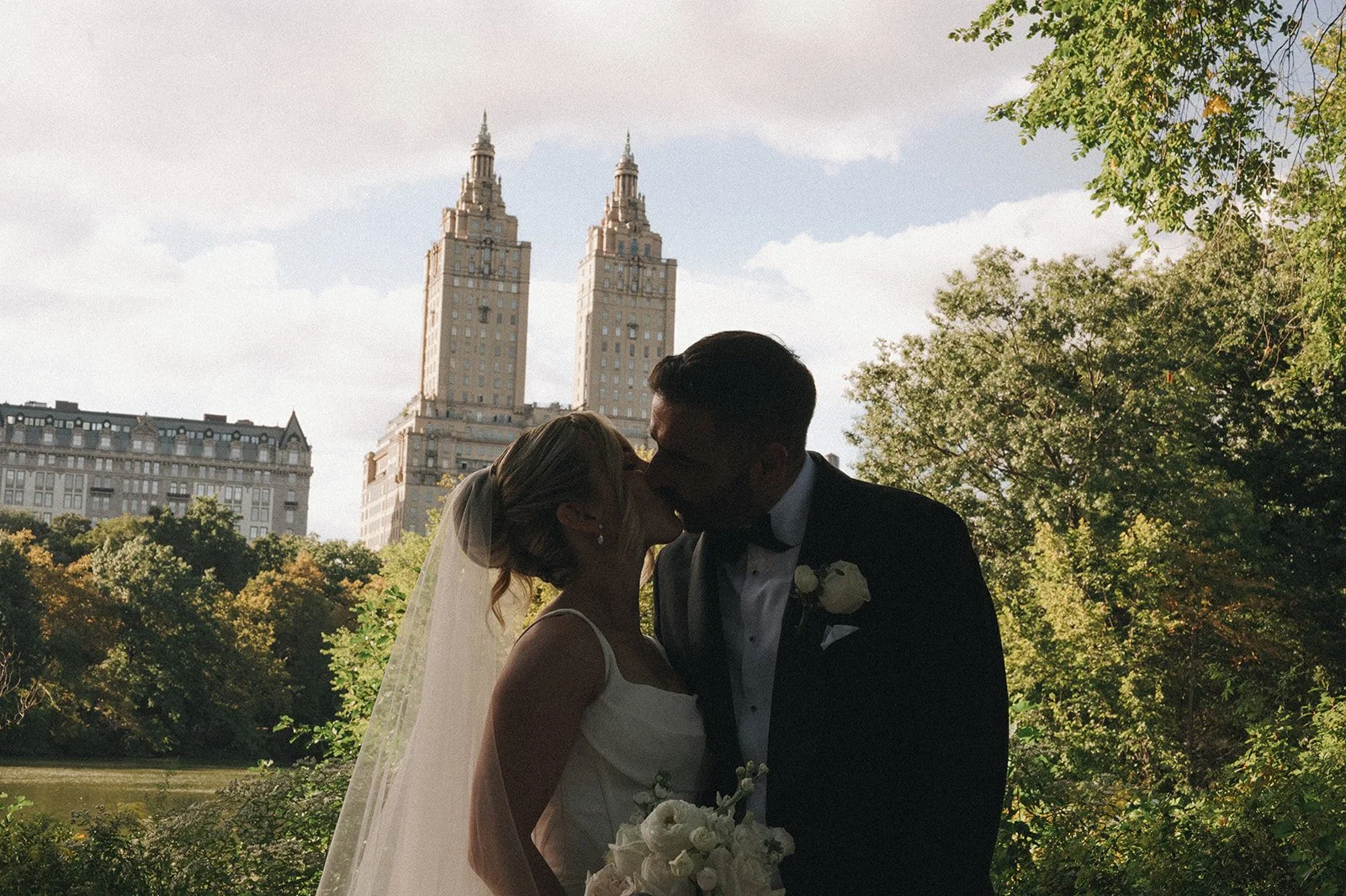 Bride and groom kissing in Central Park with the Manhattan skyline and water behind them.