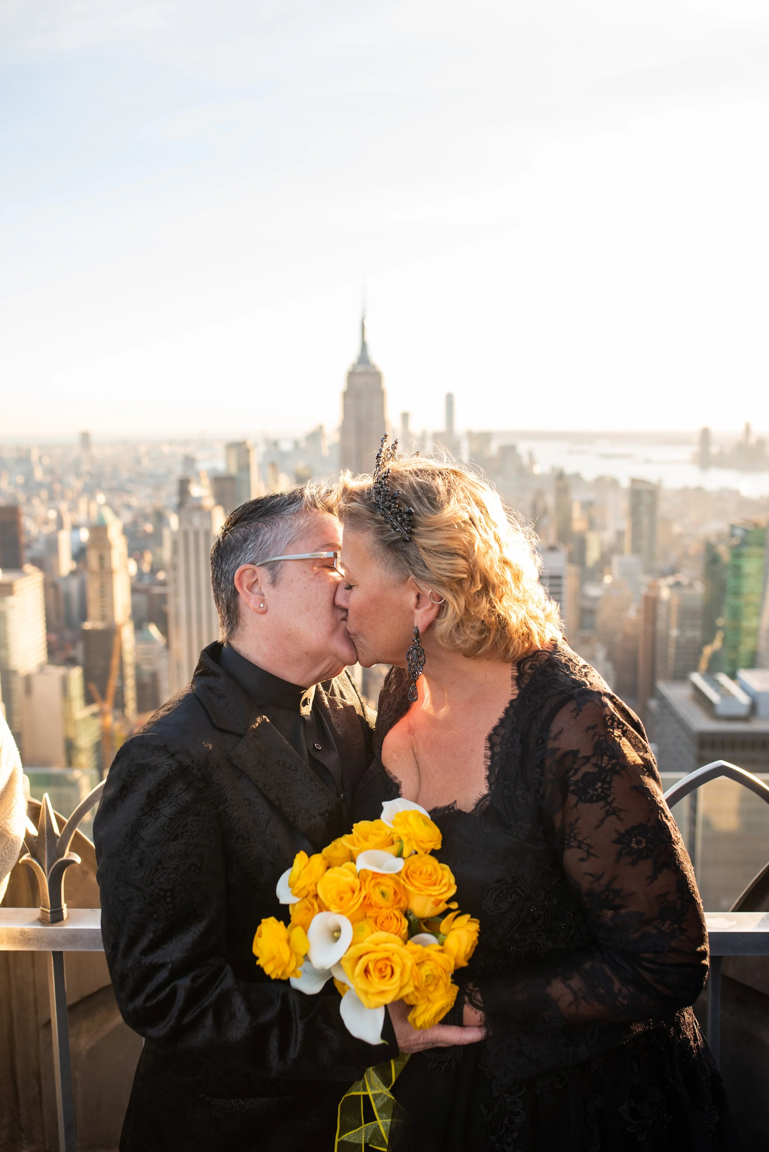 Two women wearing black and holding a yellow bouquet face kiss each other at the Top of the Rock with the Empire State Building in the background.